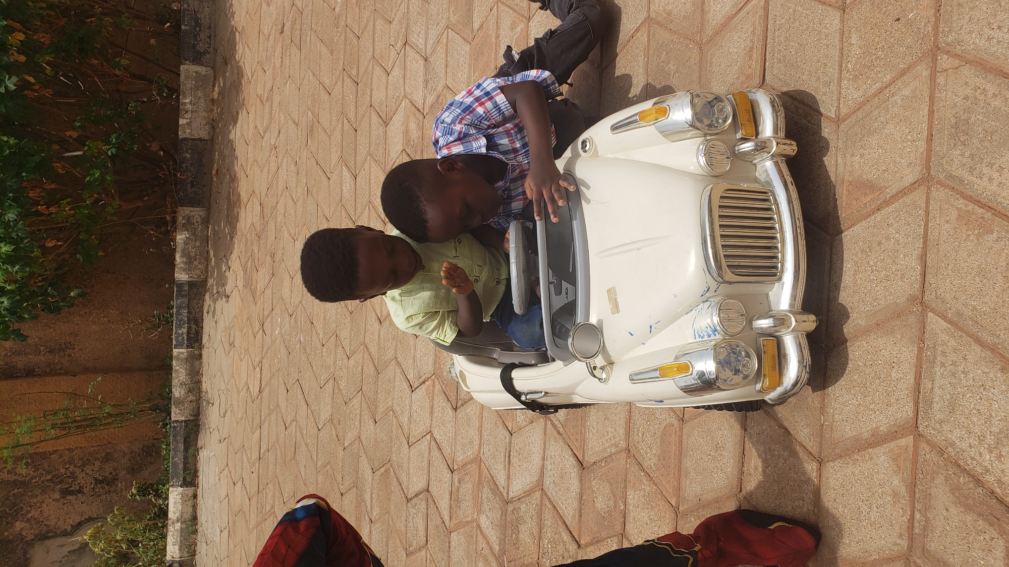 Two young Black boys are sitting in a cream-colored toy car on a paved area. The boy on the left is wearing a light green shirt and jeans, while the boy on the right is wearing a blue and red plaid shirt with black pants. They are both focused on the car's steering wheel and dashboard. The paving stones are light brown with a zigzag pattern. A concrete wall and some greenery are visible in the background. Part of a person's leg and shoe is visible at the bottom of the picture. The picture appears to have been taken outside in daylight. The city of Sabon Tasha, Nigeria, is associated with this image.