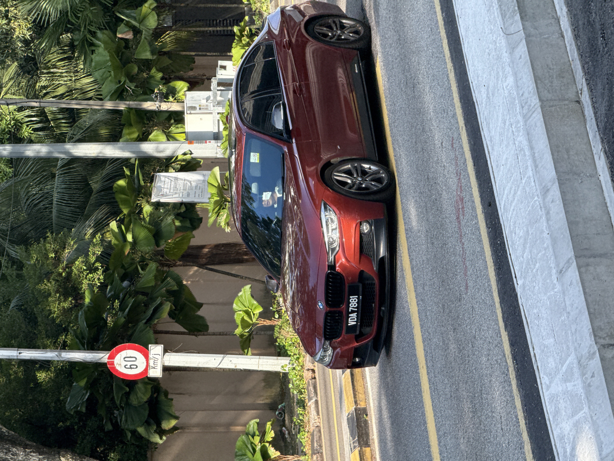 A red BMW sedan is parked on the side of a road in Kuala Lumpur, Malaysia. The car is facing toward the viewer's right. The license plate reads "VDA 7881". A speed limit sign indicating 60 km/h is visible on the left side of the frame. Lush tropical vegetation is present in the background. The road has double yellow lines. A concrete curb is to the right of the road.