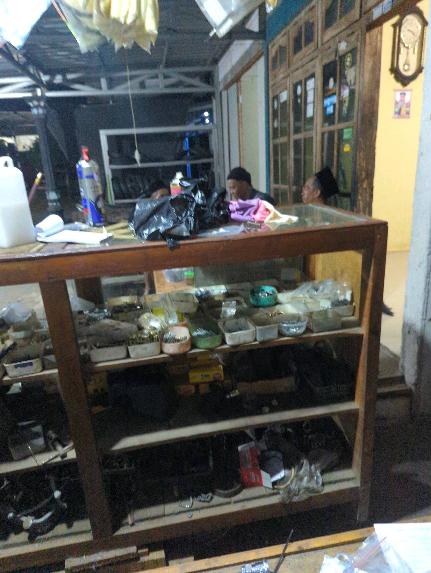 The image depicts an indoor scene, possibly a shop or a market stall, with a wooden display cabinet as the central focus. The cabinet has multiple shelves filled with various items, appearing to be trinkets, hardware, or small goods. The shelves are cluttered, with some items in containers and others loose.
In the background, two individuals are partially visible. One person, wearing a dark cap, is looking to the right. Another person, also wearing a dark cap, is mostly obscured but seems to be engaged in an activity behind the display.
The environment appears somewhat disorganized and dimly lit, with hints of a building structure in the background, including what looks like wooden doors and a yellow wall. A pendulum clock is mounted on the yellow wall. There are also plastic bags and other assorted items scattered around the area. The overall impression is of a working space or a retail establishment.