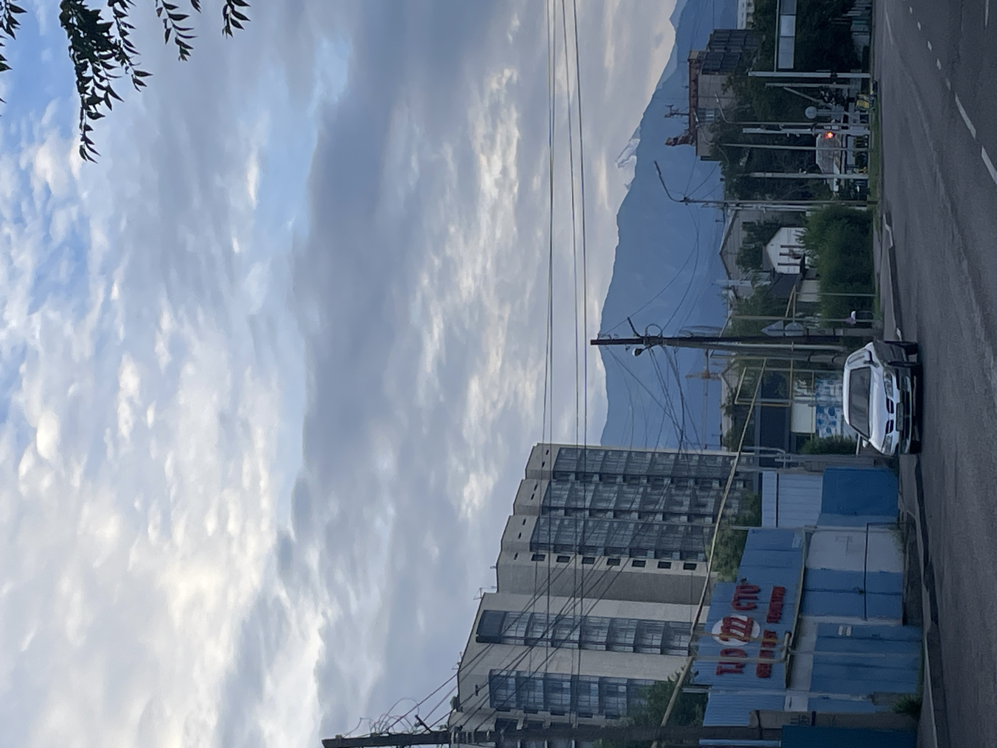 The image captures a street scene in Almaty, Kazakhstan, with a focus on urban elements and a mountainous backdrop. In the foreground, a silver sedan is parked on the right side of the road. The road itself is paved and marked with white lines. To the left of the road, a blue and white structure with red signage is visible. Further back, a large, multi-story apartment building with numerous windows dominates the mid-ground. Beyond the buildings, a mountain range, partially obscured by clouds, forms the background. The sky is overcast with dramatic cloud formations. Telephone poles and power lines are present throughout the scene, indicating urban infrastructure. The overall impression is that of a typical city street with residential buildings and natural scenery.