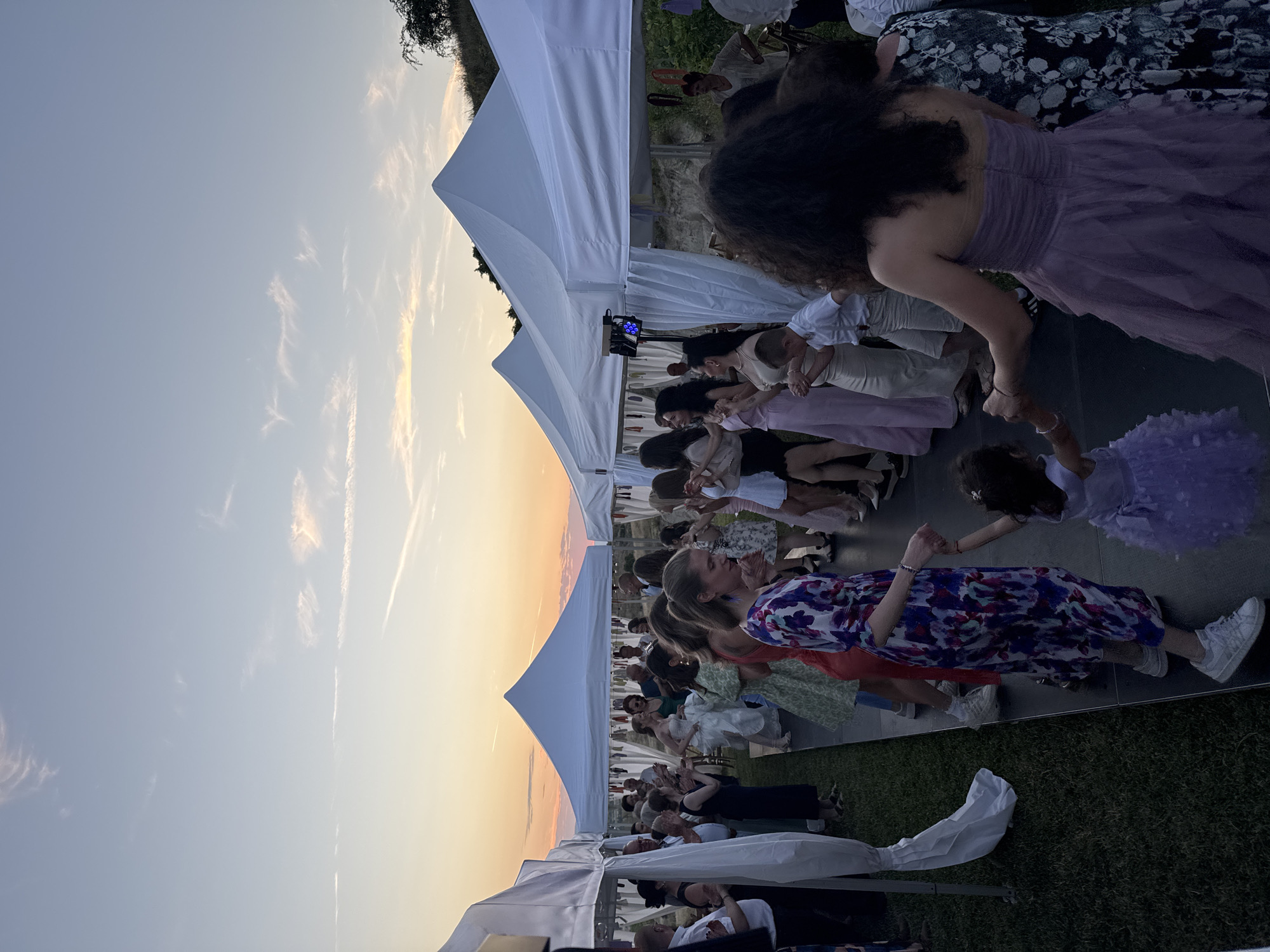 This image captures an outdoor evening event in Harsovo, Bulgaria, at sunset. The scene shows approximately 30-40 individuals, ranging from young children to adults, engaged in a social gathering or celebration. Multiple white canopy tents with draped white curtains are erected on a grassy area, with a dark, modular dance floor laid out in front of them.
The sky exhibits a clear gradient, transitioning from a light blue at the zenith to warm orange and pink hues near the horizon, with subtle cirrus clouds and faint contrails visible. To the right, a natural hill with sparse vegetation defines the background.
On the dance floor, numerous people are actively dancing. Many are holding hands, suggesting group or line dancing. In the immediate foreground, a woman with dark hair, wearing a strapless, ruffled purple dress, holds the hand of a small child in a lavender ruffled dress; both are dancing. To their left, another woman in a blue and purple floral patterned dress and white sneakers is also dancing, holding the child's other hand. Further back on the dance floor, a woman in a white dress (possibly a bride) is seen dancing. A speaker or lighting unit with blue illumination is visible under one of the tents.
To the left of the dance floor, additional guests are standing on the grass, observing the dancing and some are clapping. The overall atmosphere appears festive and interactive, characteristic of a celebratory event.