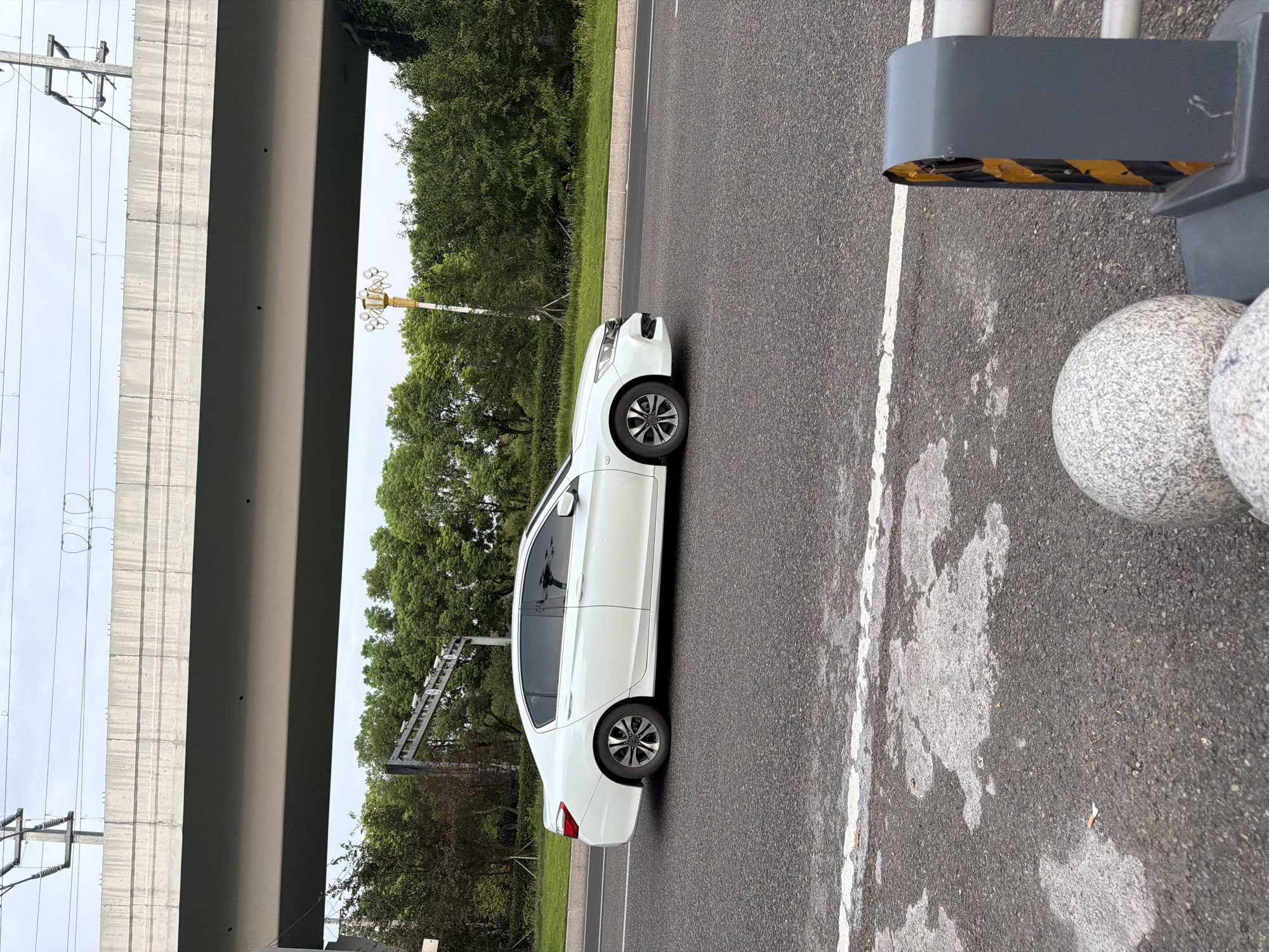 In Jiangjin, China, a white sedan is captured in profile, moving left to right on a paved road beneath an elevated highway structure. The sky is overcast. Trees and foliage border the road. The car's windows are tinted, and its wheels are visible. The road surface shows signs of wear and tear, with patches of lighter-colored material and a white lane marking. In the foreground, a section of a gray guardrail with yellow reflective stripes and two spherical stone bollards are present.