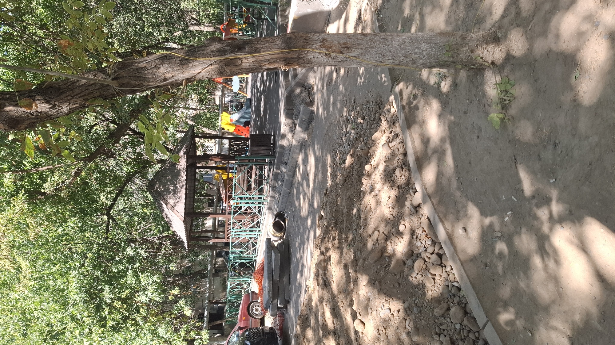 The image shows a construction site in a residential area in Shymkent, Kazakhstan. In the foreground, there is a pile of dirt and rocks, along with some concrete slabs and a pipe. Behind the construction site, there is a playground with a gazebo, slides, and swings. A red car is parked to the left of the playground. The area is surrounded by trees, indicating a possibly park-like or courtyard setting. The presence of construction materials and the partially visible playground suggest that some form of renovation or development is taking place. The time of day appears to be daytime, with sunlight casting shadows on the ground.