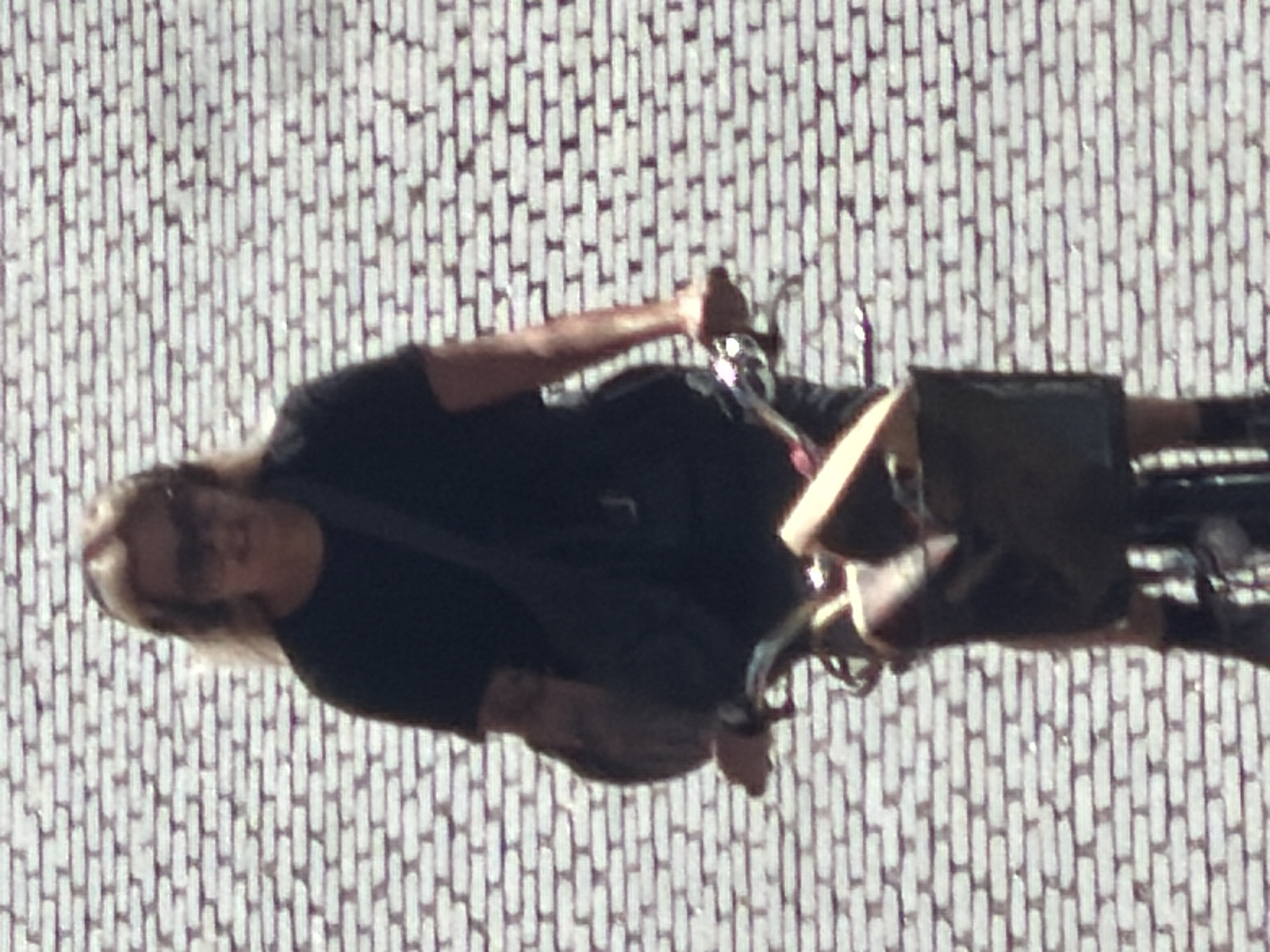 A person is riding a bicycle against a brick wall. The person has blonde hair and is wearing sunglasses and a dark shirt. The bicycle has a basket in front containing some items. The image is slightly blurry due to motion. The brick wall appears to be an outdoor urban setting. This scene is located at Folkets Park in Malmö, Sweden.