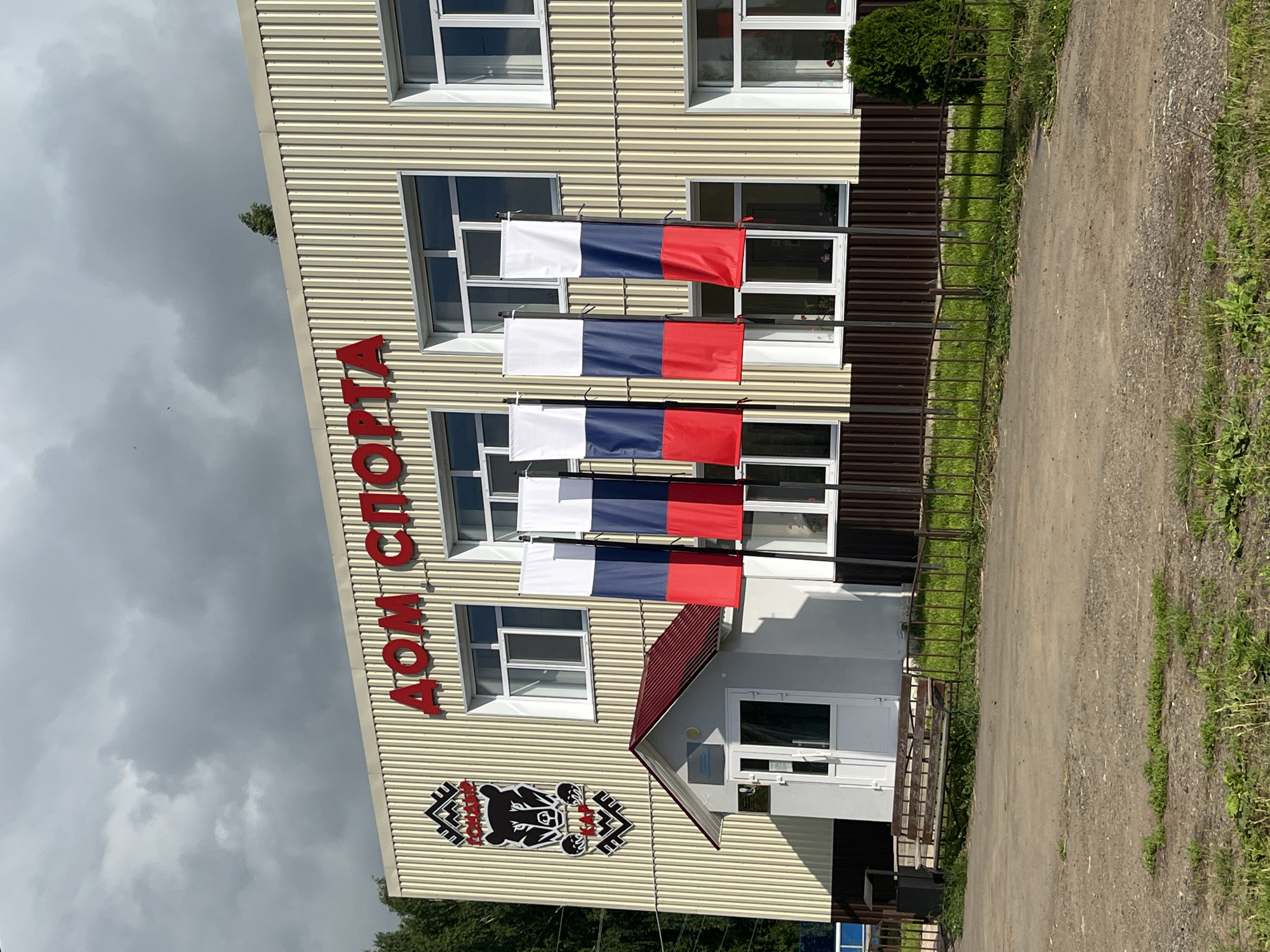 The image shows a building with the words "ДОМ СПОРТА" in large red letters. The building is beige with vertical corrugated siding. To the left of the entrance, there is a crest with a bear and some text. Three vertical Russian flags are displayed in front of the building. There is a small, white structure with a red roof to the left of the entrance, likely a ticket booth or small office. The area in front of the building is a dirt or gravel path with a small metal fence and some grass. In the background, trees are visible. The sky is overcast. The scene appears to be a sports facility.