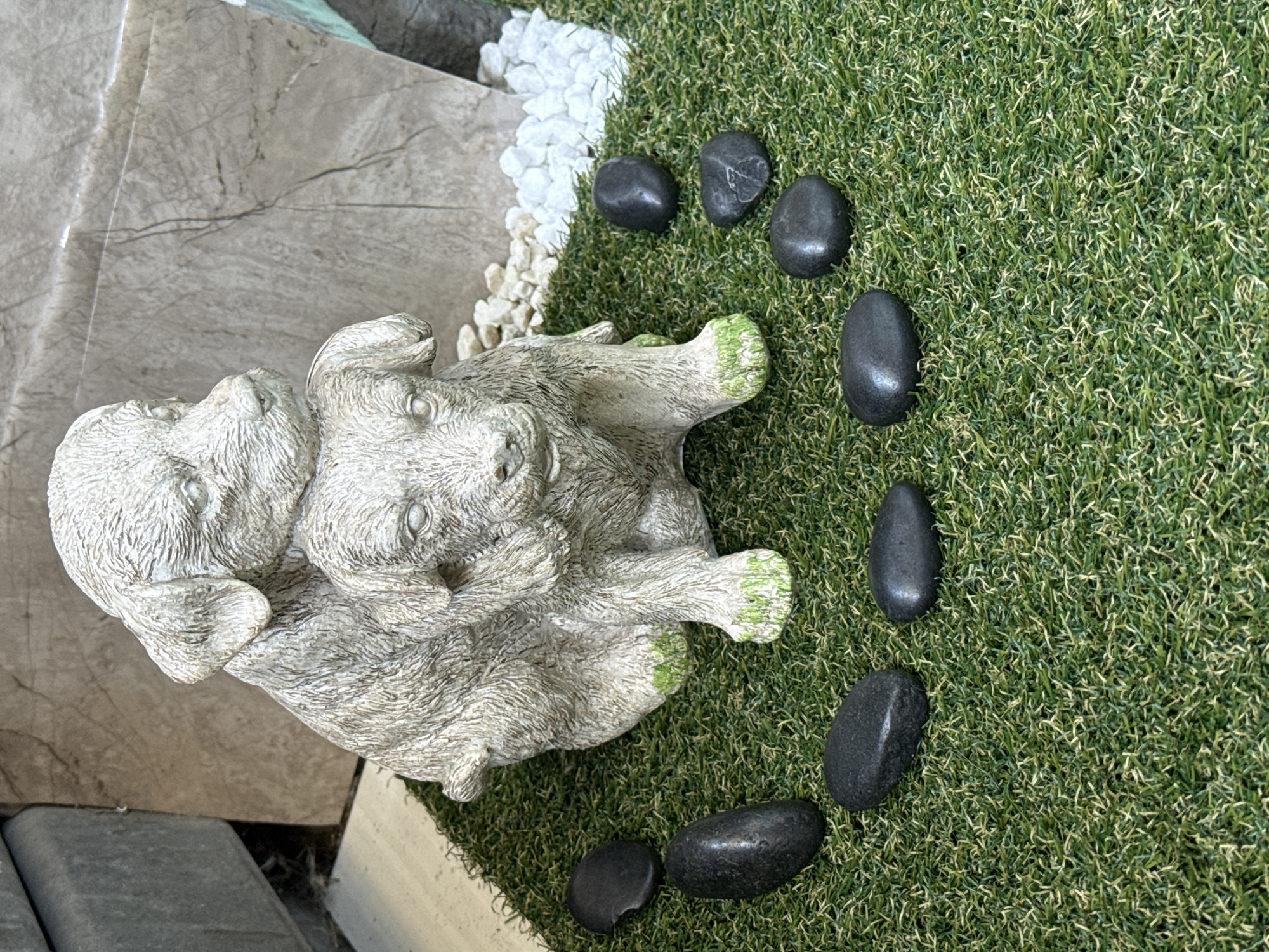 This image depicts a garden ornament featuring two dog statues, seemingly one larger dog embracing a smaller one. The statues are made of a light-colored material, possibly stone or resin, with some green moss-like growth on the paws of the lower dog. They are placed on a surface of artificial turf, which is lush and green. Scattered around the base of the statues are several dark, smooth pebbles. In the background, a large, rough-textured rock is visible, along with a cluster of smaller white pebbles. To the left of the statues, a portion of a grey, textured surface, possibly a wall or planter, is visible. The scene appears to be an outdoor decorative arrangement. There are no people or animals present. The lighting suggests it is daytime. The image has been taken from a slightly elevated angle, looking down at the arrangement.