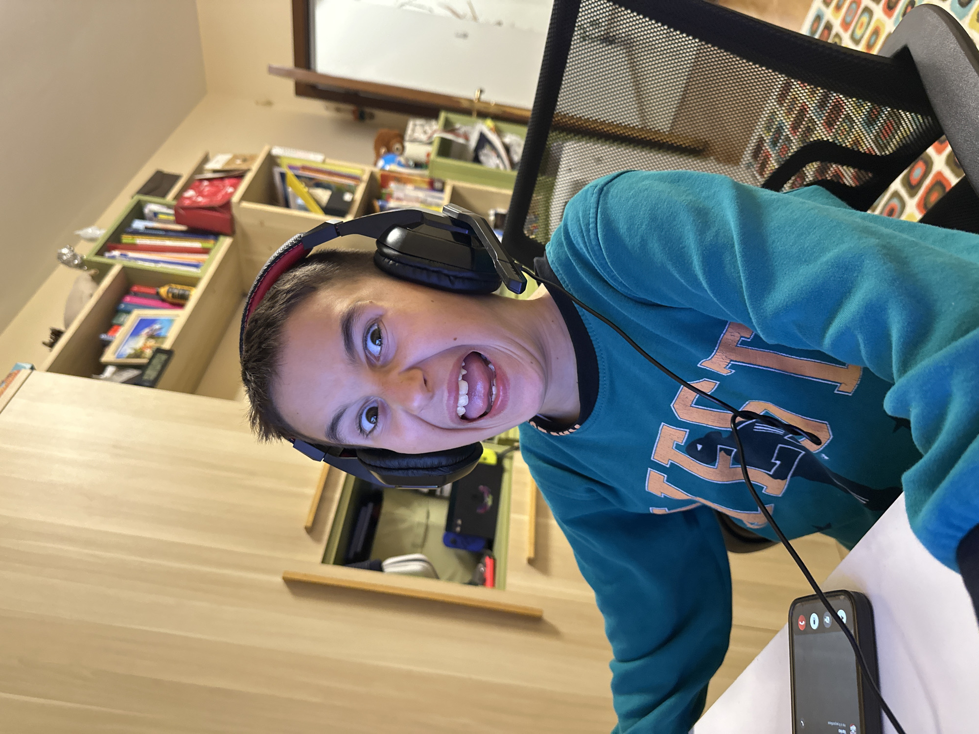 An expressive young individual, likely a child or pre-teen, is captured in a close-up, tilted portrait indoors, possibly in a child's bedroom or study in Plovdiv, Bulgaria. The subject has short dark hair and wears large black over-ear headphones, connected by a black wire to a smartphone in the foreground. They are dressed in a teal or turquoise long-sleeved shirt featuring a partially visible light orange graphic, which appears to be part of a letter or logo.
The person's face is highly animated, with wide-open eyes and an exaggerated, wide-open mouth revealing teeth and tongue, suggesting an emotion of excitement, surprise, playfulness, or intense focus.
The background reveals wooden shelving units filled with various items, including books, small boxes, a red pouch, framed pictures, and electronic devices such as a Nintendo Switch in its dock. A black mesh office chair is visible behind the person. In the foreground, on a light wooden surface, sits a black smartphone displaying an active call interface with a red hang-up button, a microphone icon, and a speaker icon. The scene captures a candid moment, likely during a gaming session, a video call, or while listening to music. The overall impression is one of youthful energy and engagement.