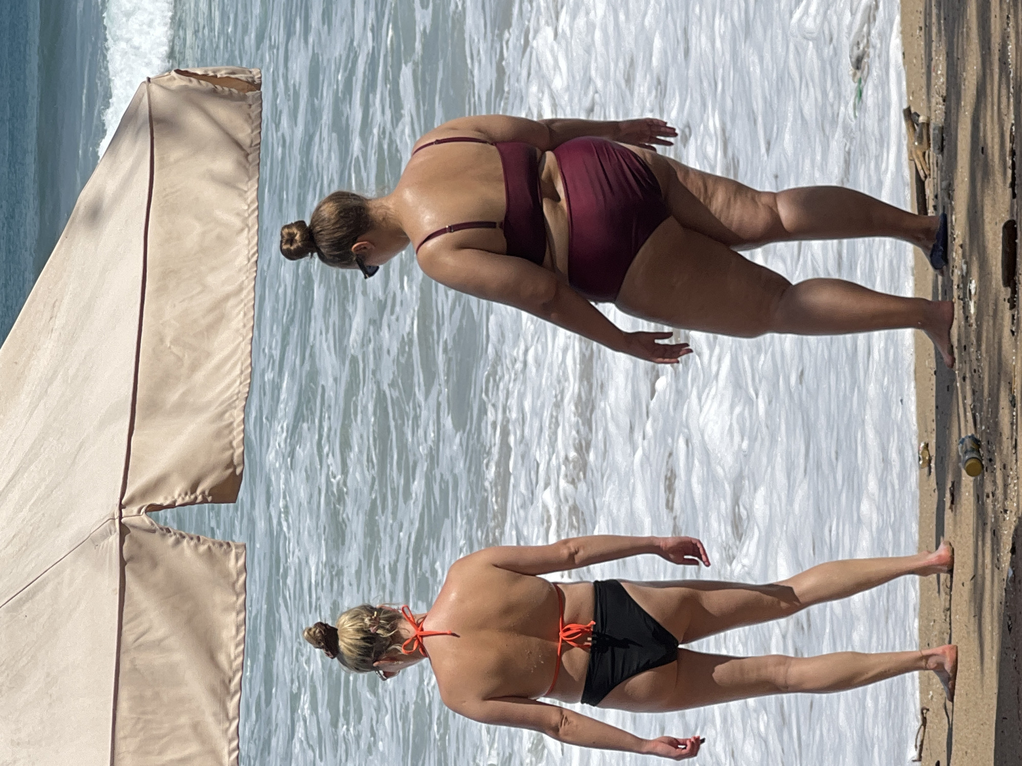 In Kuta, Indonesia, two women stand on a sandy beach facing the ocean. They are both wearing bikinis and appear to be enjoying a day at the beach under a large beige umbrella. The woman on the left has on a black bikini with an orange halter top. Her hair is pulled up in a bun. The woman on the right wears a burgundy bikini. Her hair is also styled in a bun, and she has on sunglasses. The ocean is calm with gentle waves rolling onto the shore. The beach is clean with some scattered driftwood and a discarded can.