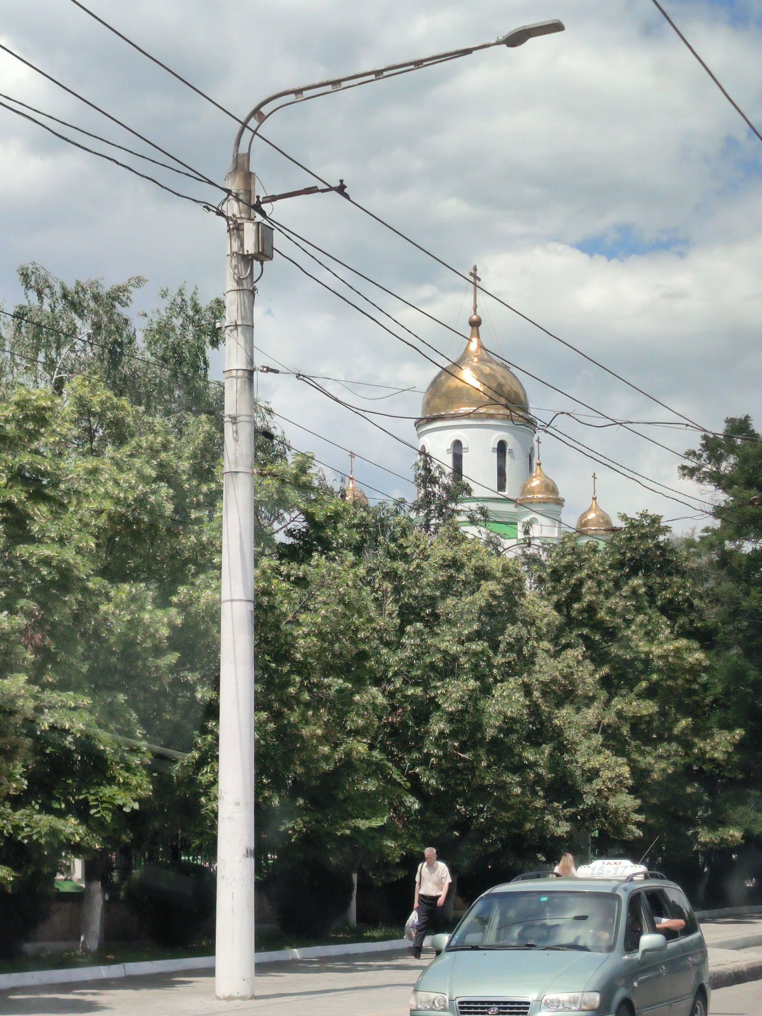The image depicts a street scene in Tiraspol, Moldova, featuring a prominent lamppost in the foreground. Behind the lamppost, a light green minivan with a "TAXI" sign on its roof is visible, driving on the road. A man in a light-colored shirt and dark pants is walking on the sidewalk to the left of the minivan, carrying a plastic bag. Further in the background, a white building with golden domes, likely a church, is partially obscured by lush green trees. The sky is overcast with scattered clouds. The environment appears to be a typical urban street with trees lining the sidewalk and road.