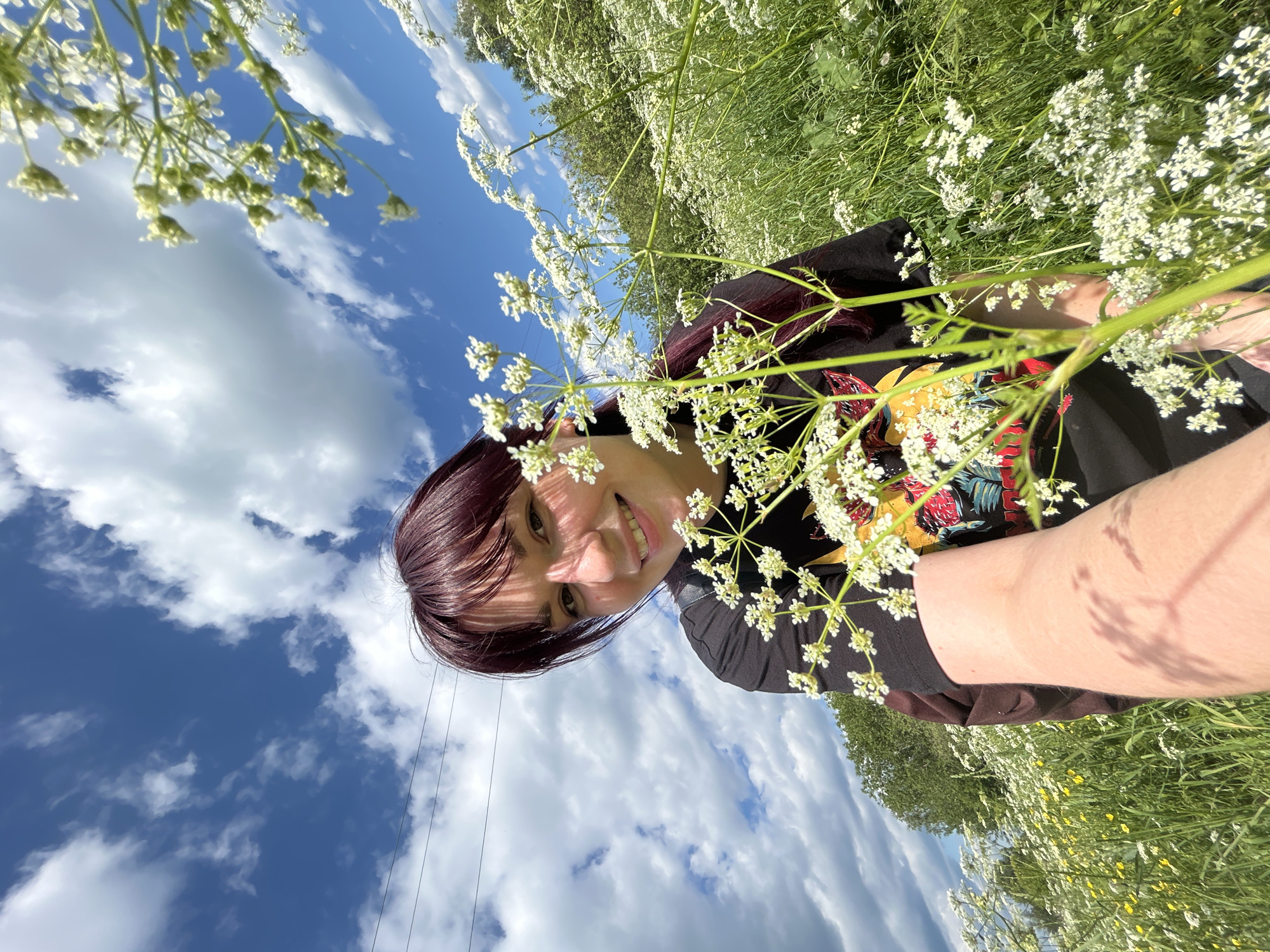 A young woman with dark hair and bangs smiles directly into the camera. She is wearing a black t-shirt with a colorful graphic design. The photo is taken outdoors, possibly in a field of wildflowers, with white flowers and green grass visible. The sky is blue with scattered white clouds, suggesting it is daytime with fair weather. The image is oriented sideways. There is no visible text.