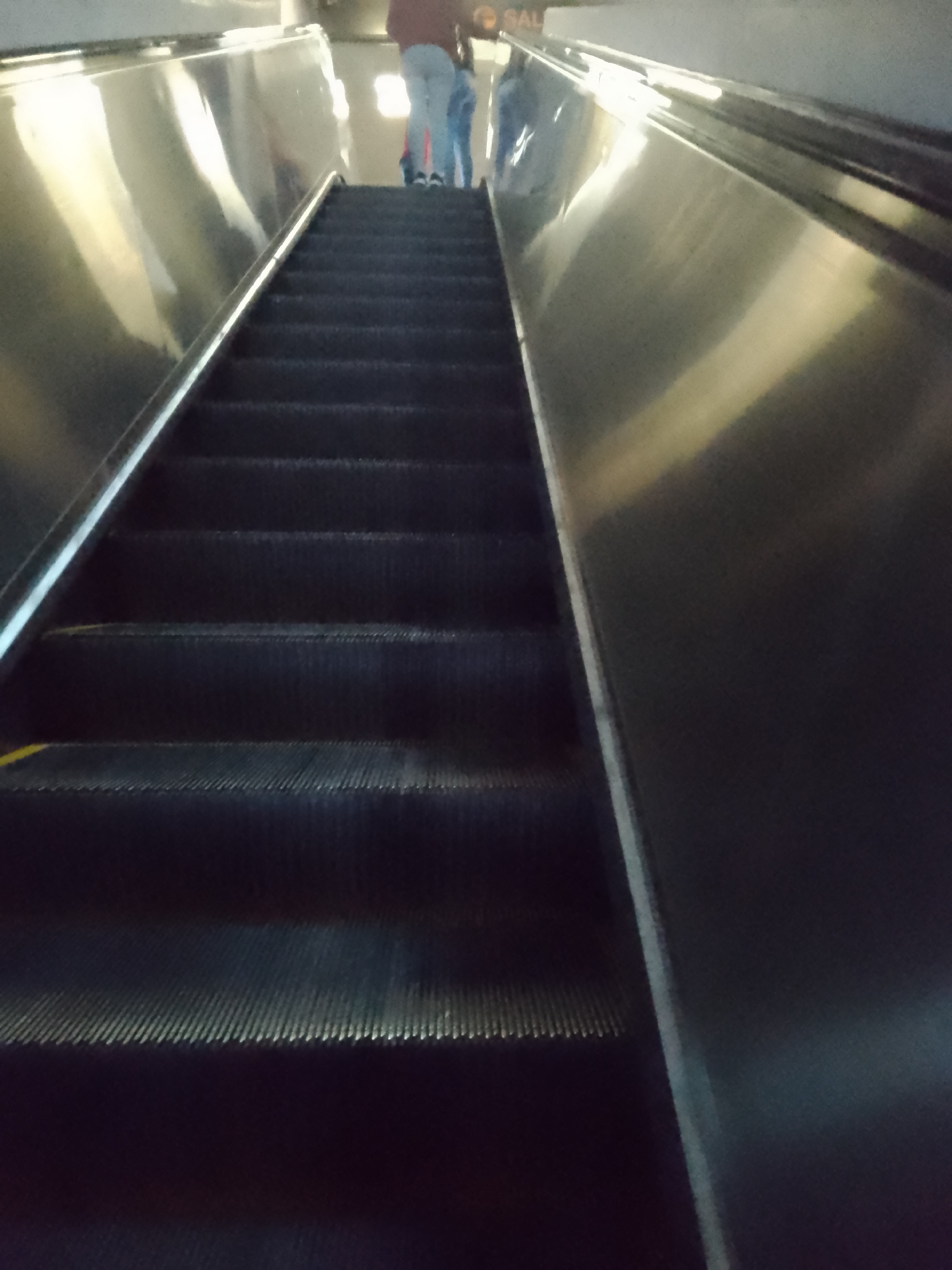 The image depicts a moving escalator ascending upward. The escalator has a dark, ribbed surface for steps and is bordered by a shiny, metallic structure on either side. Towards the top of the escalator, blurred figures of people are visible, suggesting they are either riding the escalator or standing at the top. The lighting is somewhat dim, and reflections are noticeable on the metallic surfaces of the escalator.
The location is the Caracas Metro, in the city of Caracas, Venezuela.