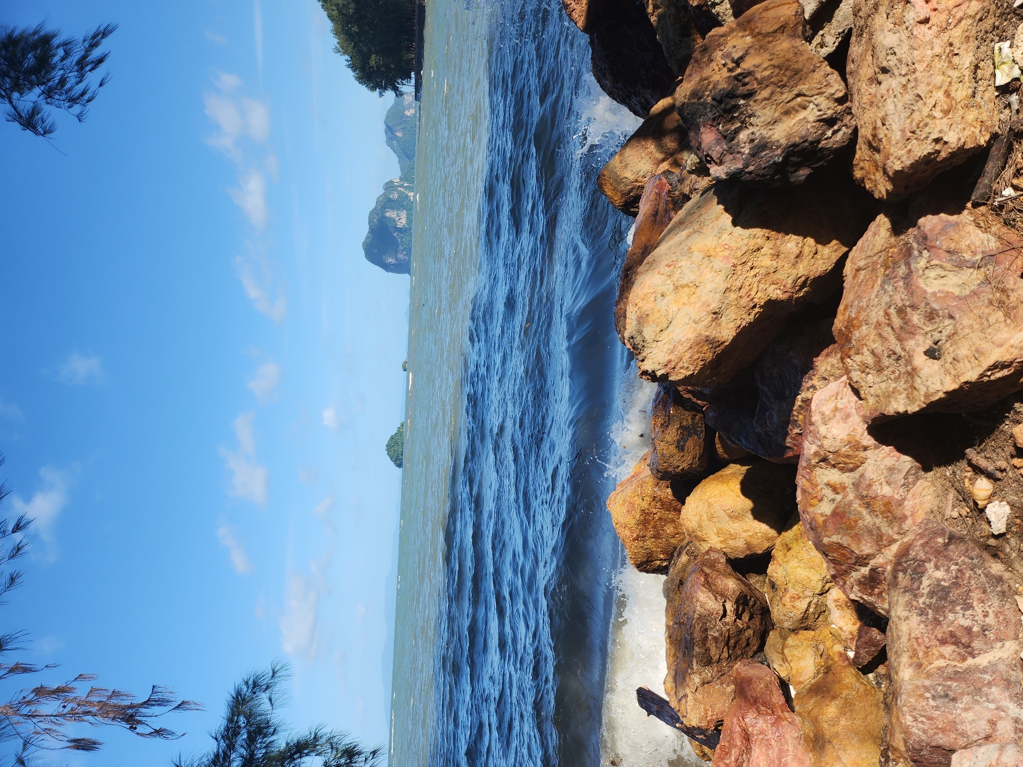 This image depicts a coastal scene at Hat Chang Lan, Thailand. The foreground is dominated by large, rough-textured rocks of various earthy tones, including brown, red, and yellow hues, some of which are wet from the sea spray. A piece of driftwood is visible among the rocks. In the mid-ground, the dark blue, choppy water of the sea crashes against the shore, creating white foam and splashes. The sea extends to the horizon, where the water appears lighter, possibly due to a change in depth or water clarity. In the distance, several islands with lush green vegetation and steep, rocky formations are visible. The sky is a clear, bright blue with a few scattered white clouds. The upper edges of the image are framed by the branches and needles of a conifer tree, with some branches appearing dry and brown. The overall impression is a rugged, natural coastal landscape under a bright sky.