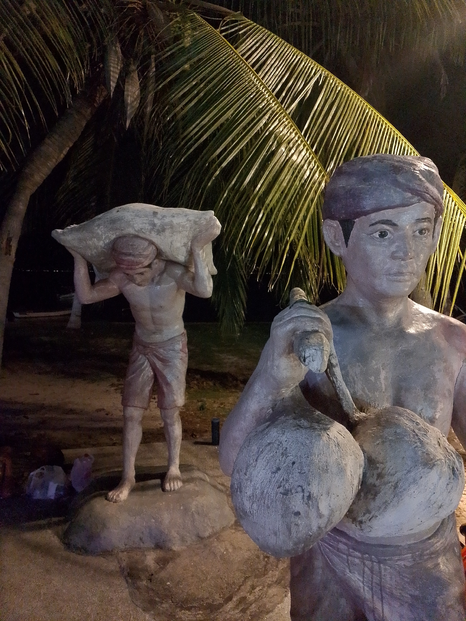 The image depicts two concrete or stone statues of male figures set against a backdrop of illuminated palm trees at night in ตำบลเกาะพะงัน, Thailand.
In the mid-ground on the left, a full-body statue of a man stands on a low, irregular base. He is depicted carrying a large, light-colored, amorphous object, possibly a sack or bundle, balanced across his shoulders. His head is bowed, and he wears a head covering and a wrap around his lower body, extending to his knees. The statue exhibits a rough, greyish texture.
In the foreground on the right, the upper torso and head of a second male statue are visible. This figure faces slightly right and holds a staff-like tool over his left shoulder. His left hand also clutches two large, spherical objects resembling coconuts, which rest against his chest. He wears a dark purple or maroon headwrap and a lower body wrap, partially visible. The statue's skin tone is a uniform light grey, with visible cracks and worn areas, and it features defined facial characteristics and a muscular build.
Behind the statues, multiple palm trees are illuminated, with their fronds displaying vibrant green and yellow hues against the dark night sky. The ground beneath the statues appears to be sand or dirt. Faint dark shapes, possibly discarded items or containers, are visible on the ground near the base of the left statue and in the lower right corner. The scene is lit by artificial light sources, casting strong highlights and shadows, particularly on the palm fronds and the textured surfaces of the statues.