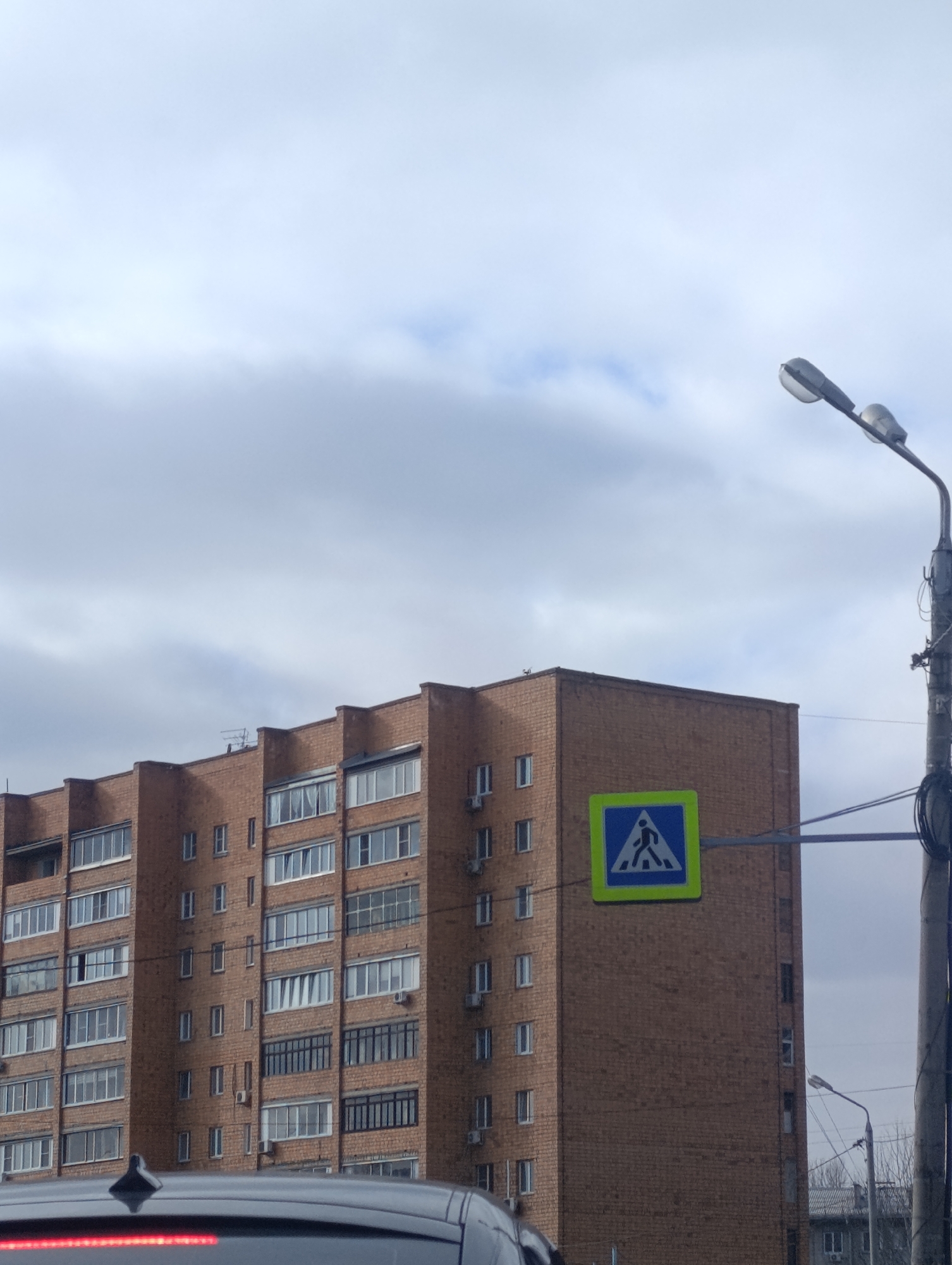 The image depicts an urban street scene featuring a large brick apartment building under an overcast sky. The multi-story building, constructed from reddish-brown brick, is a prominent subject, characterized by numerous windows, some of which have air conditioning units or enclosed balconies. Its style is typical of residential blocks found in Russian cities.
To the right of the building, a distinctive blue and yellow pedestrian crossing sign is visible. The sign is square, with a blue border framing a bright yellow inner square, which contains a black pictogram of a walking person. This sign is mounted on an arm extending from a grey utility pole, which also supports a street light fixture angled upwards and to the left, as well as an array of black cables.
In the lower foreground, the top part of a dark vehicle, likely a car or SUV, is visible, including a red light strip across its rear and a small antenna on its roof. The sky is largely covered with grey and white clouds, suggesting an overcast day with no direct sunlight. In the distance, at the bottom right, other buildings and bare trees can be faintly seen. The scene captures a moment from an ordinary day in a city like Krasnoyarsk, Russia, with no specific activity or individuals prominently featured, but with clear infrastructure for both pedestrians and vehicles.