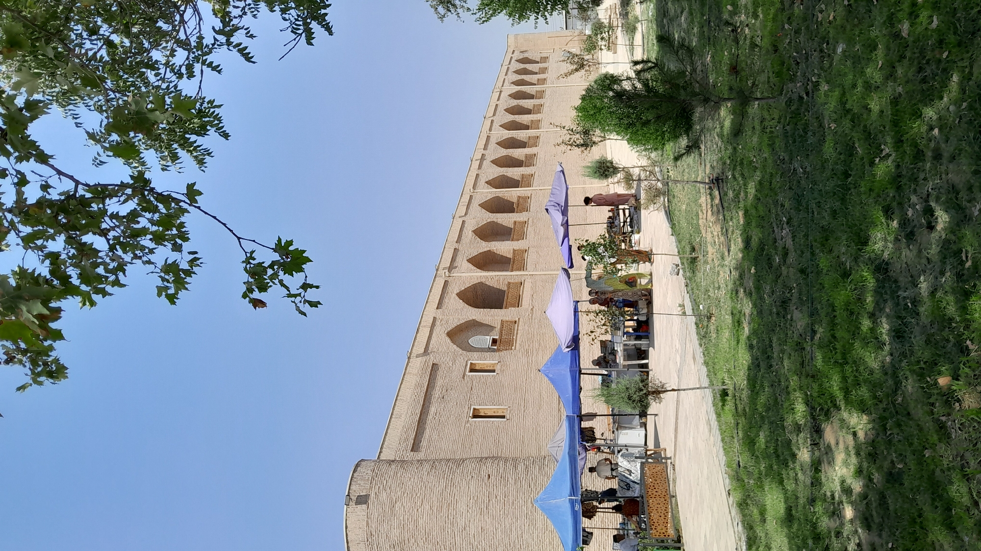 The image depicts an outdoor marketplace in Denov, Uzbekistan, in front of a long, tan brick building with arched openings. Several vendors are set up under blue and purple tents, displaying goods that appear to include books and clothing. Some people are browsing or interacting with vendors, while others are walking through the market area. The ground is paved in some sections and covered with grass in others, with several small trees and bushes scattered around. Overhead, the branches of a large tree with green leaves partially obscure the clear blue sky. The building itself features a rounded tower on the left side and numerous arched alcoves running along its length, suggesting historical architecture. The overall scene is one of a bustling daytime market in a location with both traditional and natural elements.