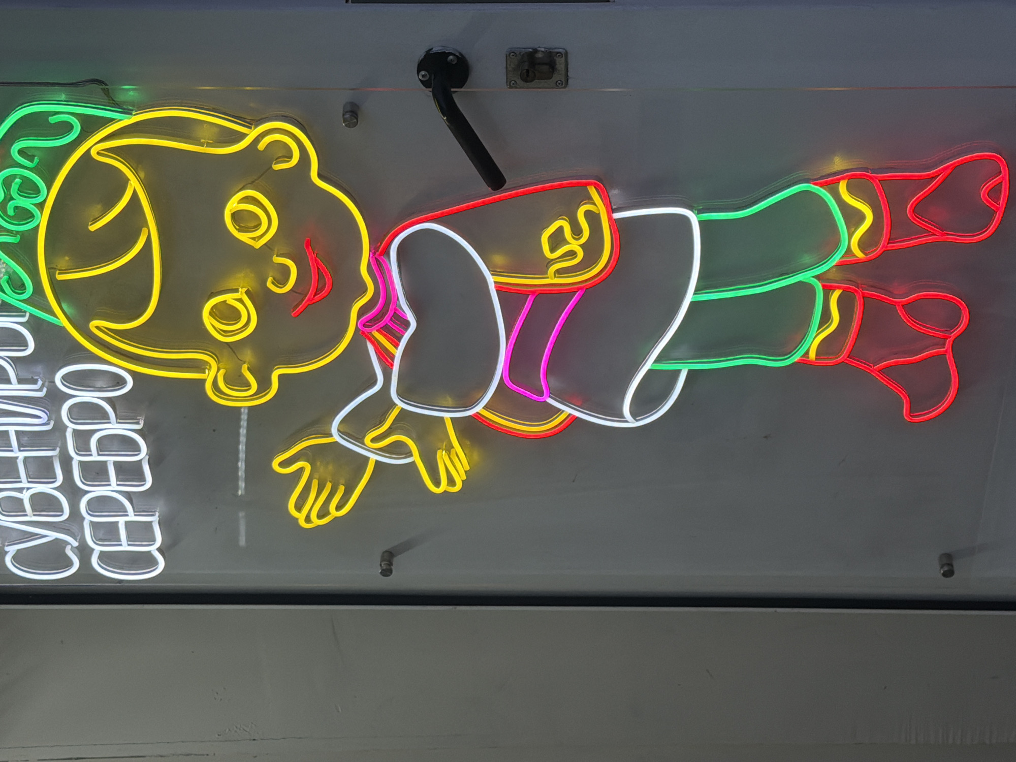 A neon sign depicting a cartoon child is mounted on a glass door in an unknown city. The sign features the child in a traditional outfit with a green cap, yellow hair, yellow eyes, and red lips. The child's clothing is outlined in white, with pink and yellow accents on the torso and green on the pants. The child's boots are outlined in red with yellow accents. Above the child's head, in white neon letters, are the words "СУВЕНИРО" and "СЕРЕБРО". The glass door has a black door handle on the right side and a metal latch below it. The background is a plain grey wall.