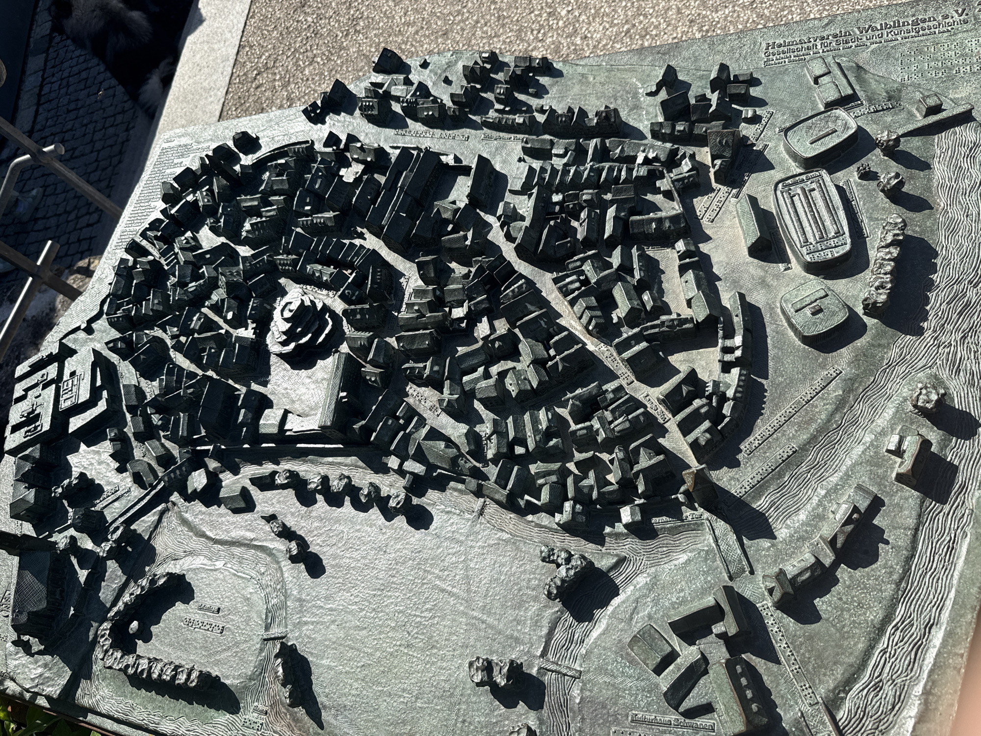 A detailed, elevated, tactile bronze or patinated metal model depicts the historical layout of Waiblingen, Germany, circa 1800. The model features a dense urban core with numerous three-dimensional building blocks, intricate street patterns, a winding river (likely the Rems) with textured water, and surrounding terrain including a fortified wall section. Prominent structures include a central, multi-tiered circular building (Stadtkirche St. Michael) within the dense city, and various larger buildings on the periphery. Labels inscribed on the model, primarily in German, identify specific locations such as "Marktplatz," "Schmiedgasse," and "Landhaus Sarazen." Informational text on the bottom right corner of the model credits "Heimatverein Waiblingen e.V." and specifies "historische Lage Waiblingen um 1800" (historical location of Waiblingen around 1800). Tactile dots are incorporated along streets and next to labels, suggesting accessibility for the visually impaired. The model is situated outdoors on a light-colored, textured paved surface, likely concrete, under direct sunlight, casting strong, defined shadows from the buildings onto the model's surface. A metal railing and a dark, indistinct object are visible in the background to the upper right.