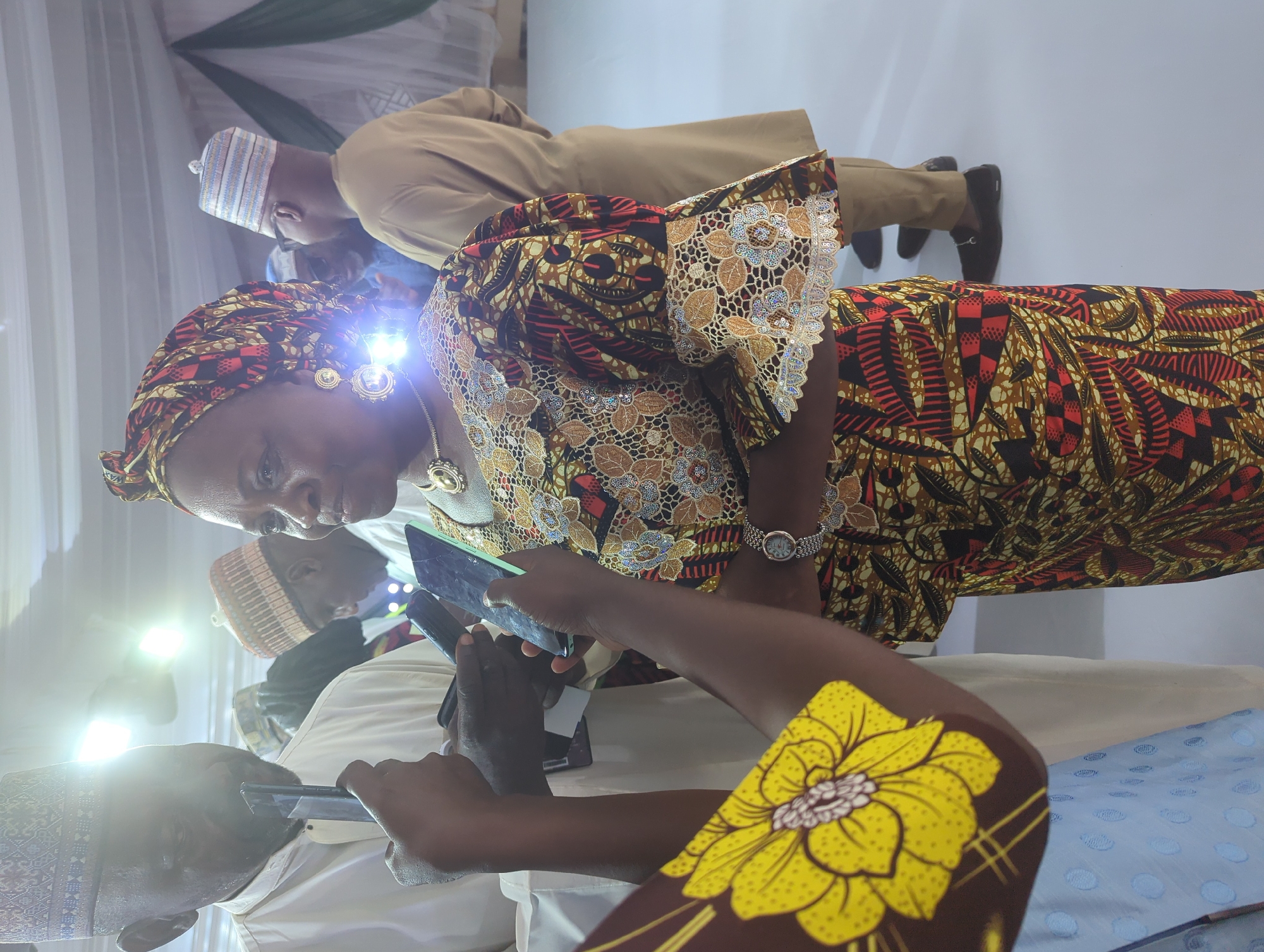 The image shows a group of people indoors, likely at an event in Unguwar Sarki, Nigeria. The orientation of the image is portrait, and it appears to be taken during the daytime. The lighting is bright, suggesting artificial lighting from within the venue, possibly spotlights as indicated by a bright glare on one of the subjects.
The main focus is on a woman in the foreground wearing a colorful, patterned headwrap and a dress with intricate lace detailing on the sleeves. She is looking down at a phone she is holding. To her right, another person's arm is visible, also holding a phone. Behind them, several other individuals are present. One man is wearing a traditional cap and a beige outfit. Another person, partially obscured, is also wearing a cap. The background consists of white and green draped fabric, possibly decorations for the event. A bright yellow fabric with a floral pattern is prominent in the lower right portion of the image.
The people appear engaged, with at least two individuals focused on their mobile phones, suggesting they might be documenting the event, communicating, or looking up information. The overall mood seems to be one of casual gathering or observance. There is no visible text in the image.