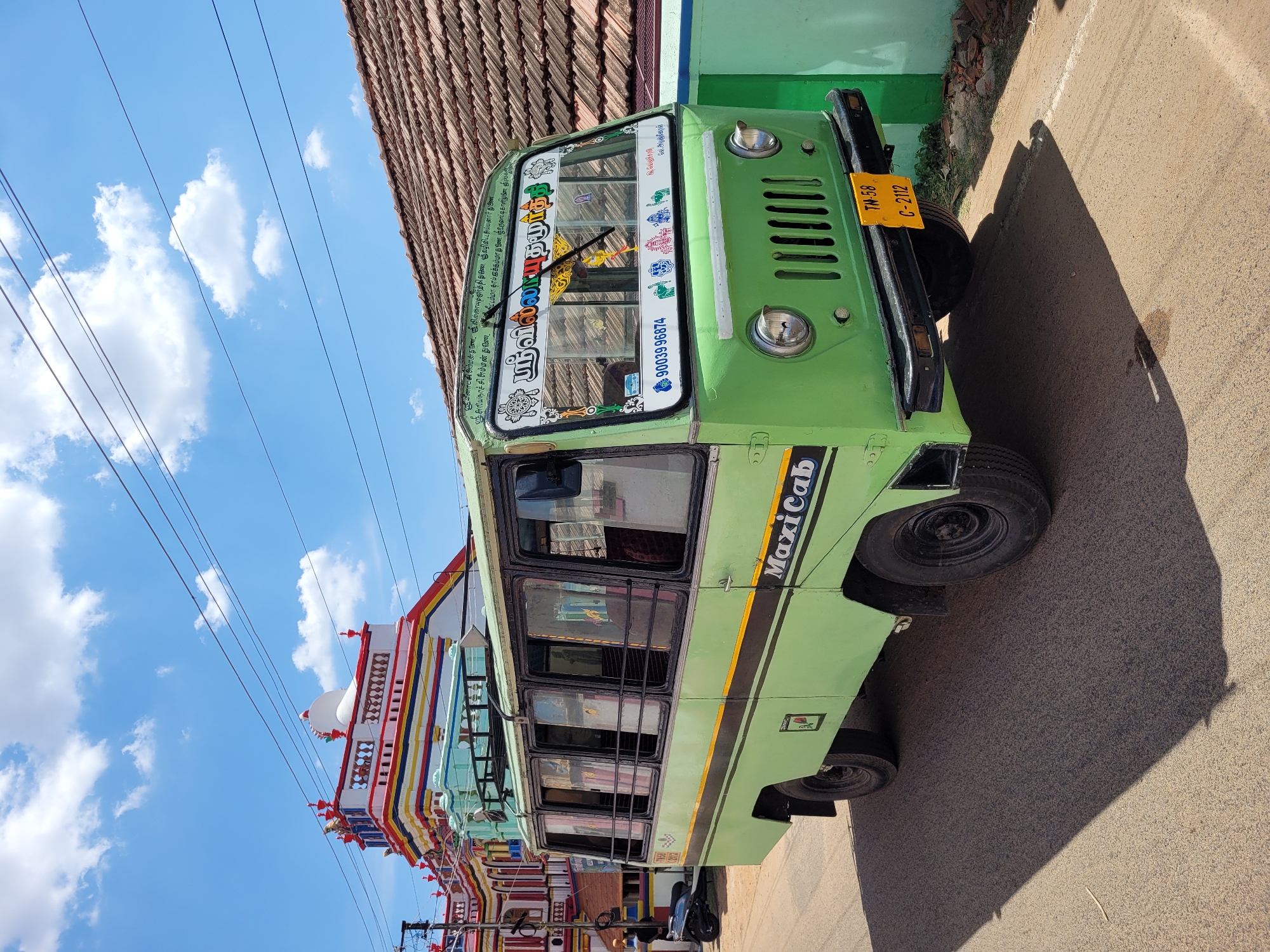 A bright green MaxiCab is parked on a sunlit street, captured in a portrait orientation that makes it appear tilted. The vehicle, which resembles a small bus or large van, features "MaxiCab" written in black capital letters on its side. Its front windshield is adorned with various decorations and Tamil script.
Prominently displayed on the windshield is the name "ஸ்ரீவில்லாயதமுர்த்தி" (Sri Villayathamurthy). Above this, smaller Tamil text invokes several deities: "ஸ்ரீ மீனாட்சி அம்மன் துணை", "ஸ்ரீ லில்ணையுமூர்த்தி துணை", "ஸ்ரீ வழிவிட்ட சிய்யனார் துணை", "ஸ்ரீ அரியநாச்சி அம்மன் துணை", "ரேடம்மா தாம் இக்கம்மாது இணை பஉருவாட்டி காளிதுகினா ஸ்ரீ" (invoking Meenakshi Amman, Lellayathamoorthy, Vazhivitta Siyyanar, Ariyanachi Amman, Redamma, Ikkamma, and Urvatti Kali). A contact number, "9003996874", is also visible on the windshield. The vehicle's yellow license plate, located on the front bumper, reads "TN-58 C-2112", indicating its registration in Tamil Nadu, India.
The bus is parked alongside a wall that transitions from green paint to an exposed tiled roof. On the opposite side, colorful, intricately decorated buildings, possibly temples or traditional houses, are visible. Overhead, several power lines stretch across a clear blue sky dotted with fluffy white clouds. Strong shadows are cast on the asphalt road, suggesting it is daytime with bright sunlight. No people are visible in the image.