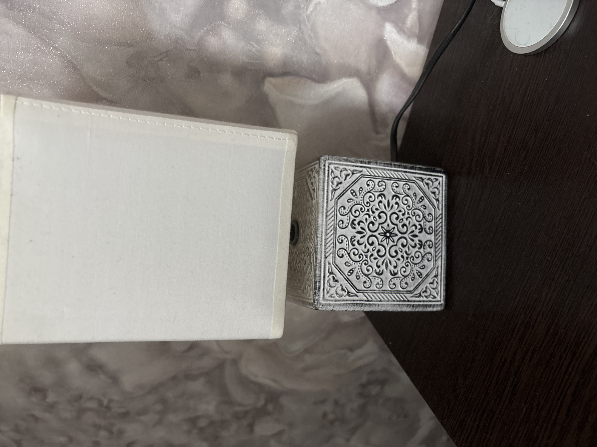 A close-up, eye-level shot shows a small table lamp on a dark wood surface. The lamp has a white fabric lampshade and a square base with an intricate black and white patterned design, resembling traditional tilework. A black power cord extends from the base and disappears to the right. In the background, a textured wallpaper with a floral motif is visible. The overall impression is one of a domestic setting, possibly a bedroom or living room, in Kinel'-Cherkasy, Russia.