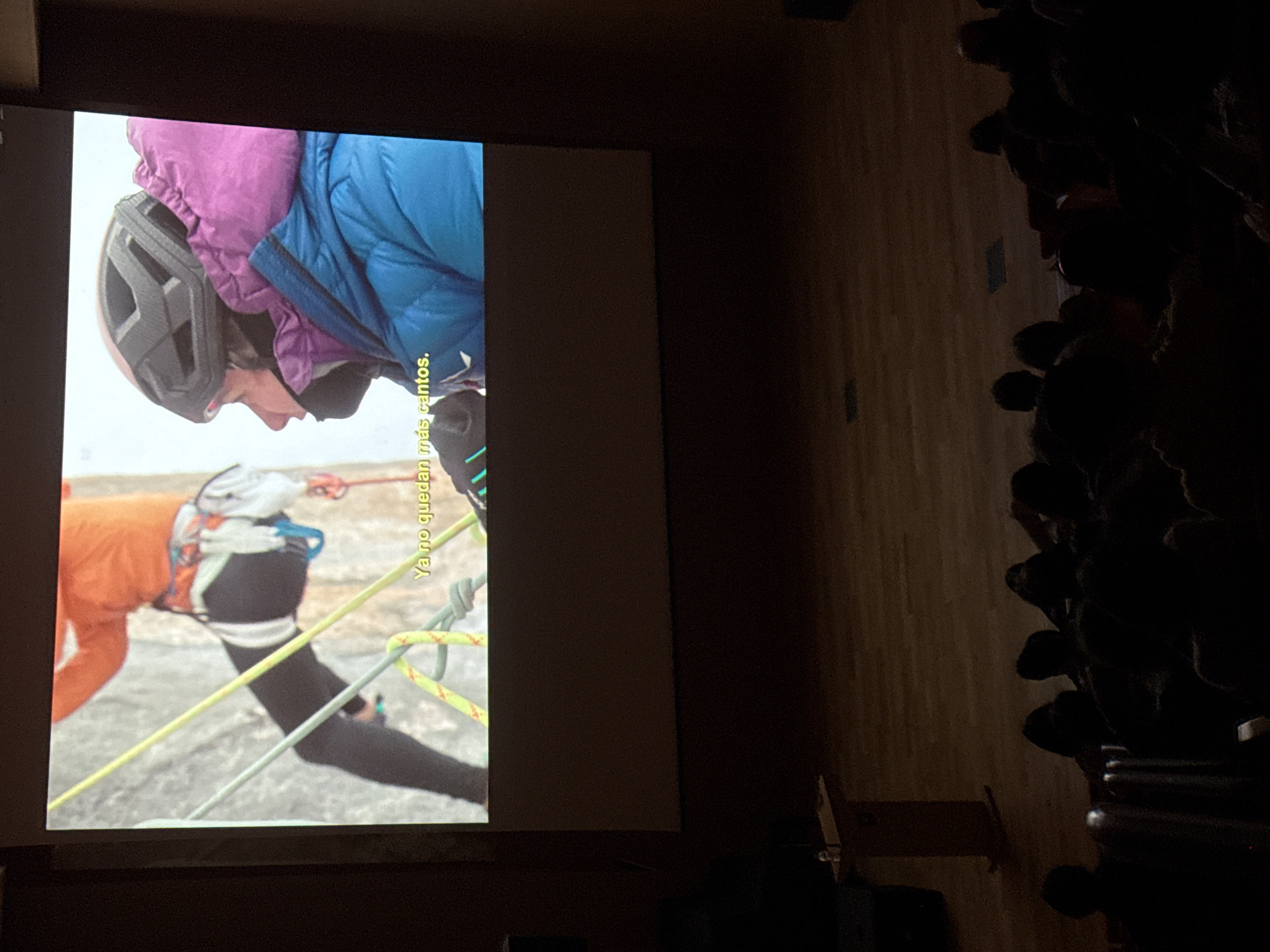 A presentation screen in a dim room shows a scene of what appears to be a mountain climber in action, wearing protective gear including a helmet, an orange top, and black climbing pants. Climbing ropes and harnesses are visible. Subtitles in Spanish, stating "Ya no quedan más cantos" or its equivalent in English, are superimposed on the scene. In the room with the screen, there are visible audience members sitting on rows of chairs. The image was taken inside a room, and no information is available to identify if it happened in Benasque, Spain, or about the relationships or intentions among the audience.