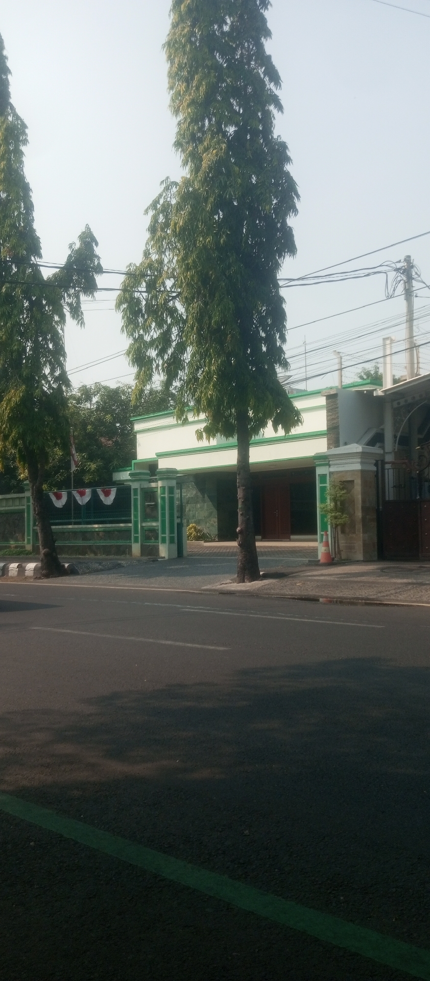 The image depicts a street scene in Kediri City, Indonesia, likely in the morning or late afternoon due to the shadows. A paved road occupies the foreground, with a green painted line marking its edge. Across the road, a low-rise building with a white facade and green trim is visible, featuring columns and a fence. Two tall, slender trees with dense green foliage stand prominently in front of the building. The background includes power lines and a hazy sky. No people are present in the image.
