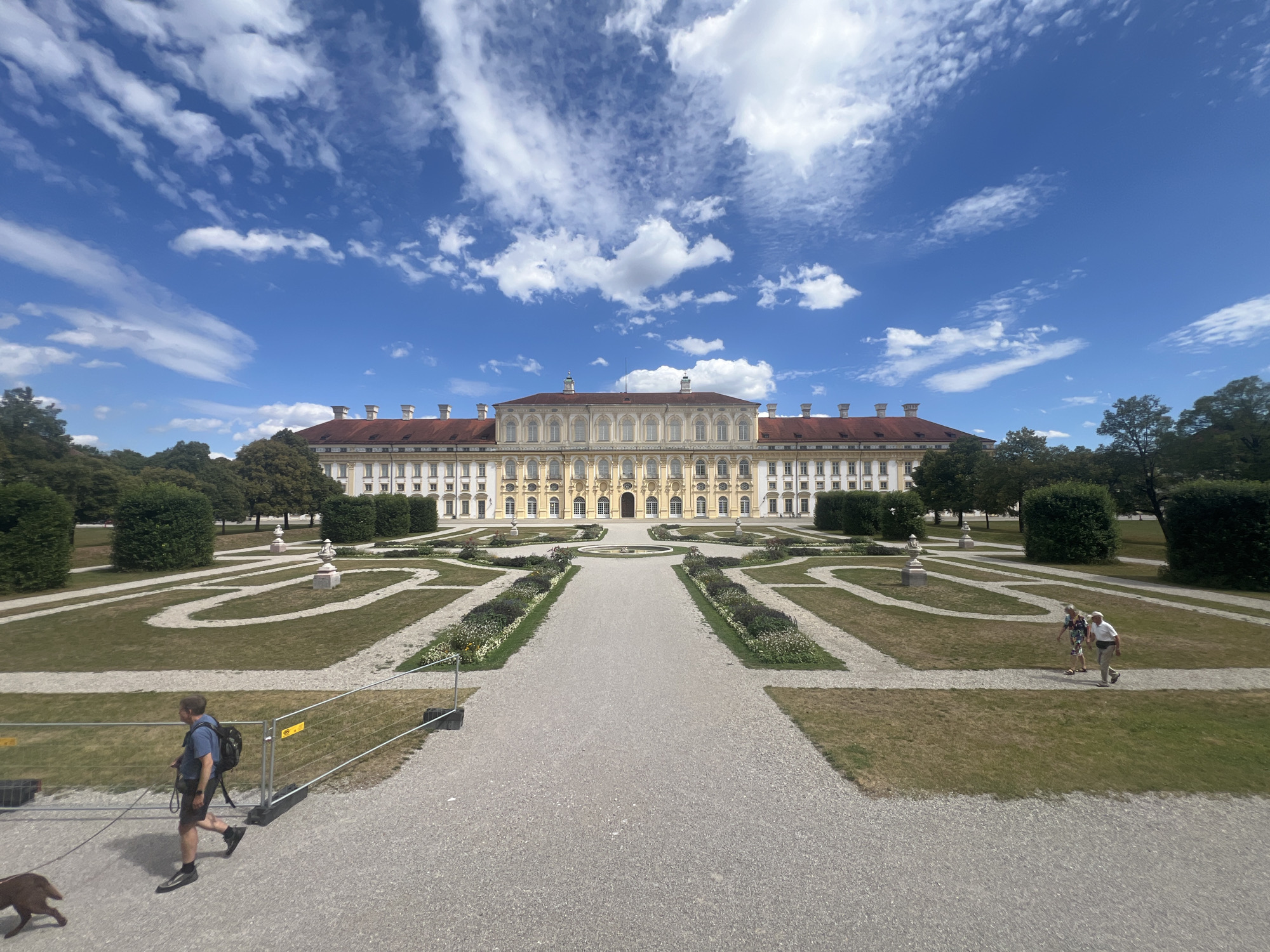 The image shows a grand baroque palace with symmetrical gardens in Oberschleißheim, Germany. The palace is a long, multi-story building with a light yellow facade, red tiled roofs, and numerous windows. It is flanked by trees and manicured hedges. In front of the palace, a formal garden features gravel pathways, manicured lawns, flower beds, and decorative statues. Two elderly individuals, a man and a woman, walk along a garden path on the right side of the image, both facing left. On the left side, a man with a backpack walks his dog on a leash along a gravel path. The sky is a vibrant blue with scattered white clouds, indicating a clear day. The overall scene depicts a well-maintained historical landmark and its surrounding parkland, with visitors enjoying the surroundings.