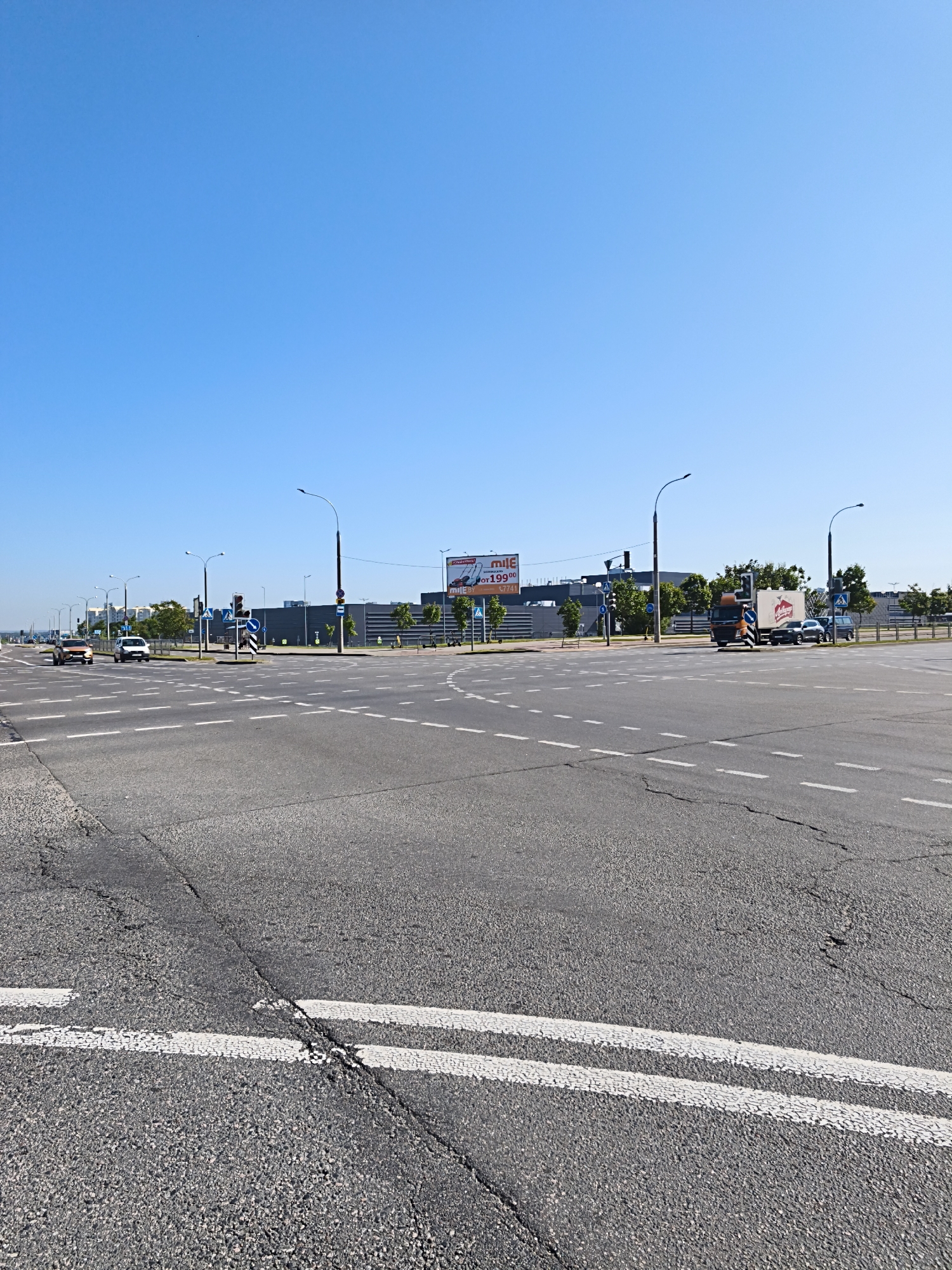 This is a wide-angle, eye-level photograph of a large intersection in Minsk, Belarus, under a clear blue sky. The asphalt road surface is marked with numerous white dashed lines indicating lanes and pedestrian crossings. In the foreground, the road appears somewhat cracked and worn.
Several vehicles are present. On the left, two cars, a silver sedan and an orange SUV, are driving away from the viewer. In the middle ground, a large truck with an orange cab and a white trailer is stopped at the intersection. To the right of the truck, a dark-colored car and a blue SUV are also visible.
Streetlights are positioned throughout the intersection, some with traffic signals. Pedestrian crossing signs, featuring the universally recognized white figure on a blue background, are visible on several corners. Behind the intersection, a long, grey, modern building with a dark facade dominates the background. To the right of this building, a large billboard advertising "Mile" is mounted. Some trees and smaller buildings are visible further in the distance. The overall scene depicts a typical urban environment on a sunny day.