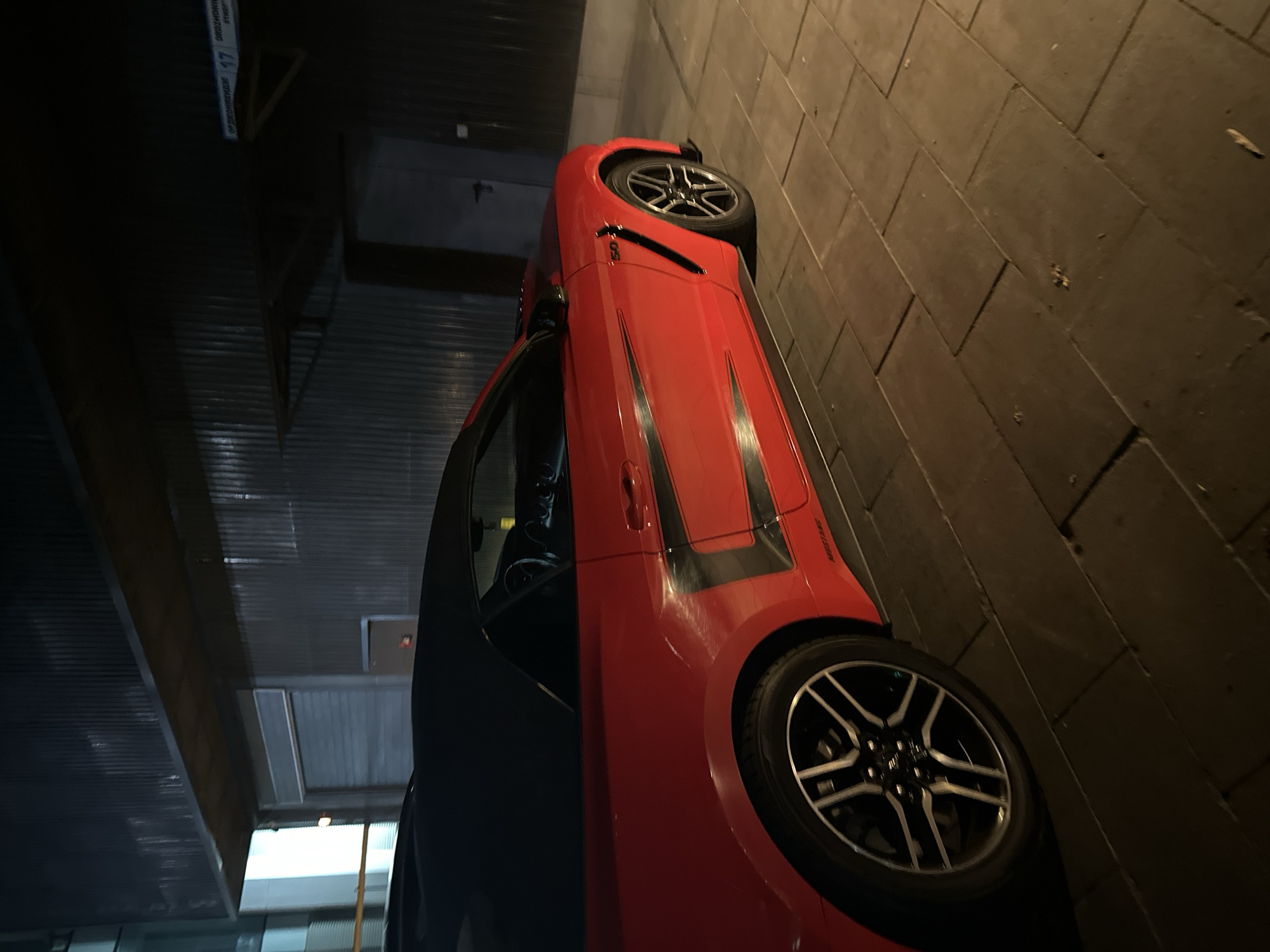 A red convertible Ford Mustang is parked outdoors at night on a paved surface. The car has black accents and a black soft top. The background consists of a corrugated metal building with a door and a sign that reads "ДЗЕРЖИНСКОГО 17" (Dzerzhinsky Street 17). The overall lighting suggests an urban environment at night.