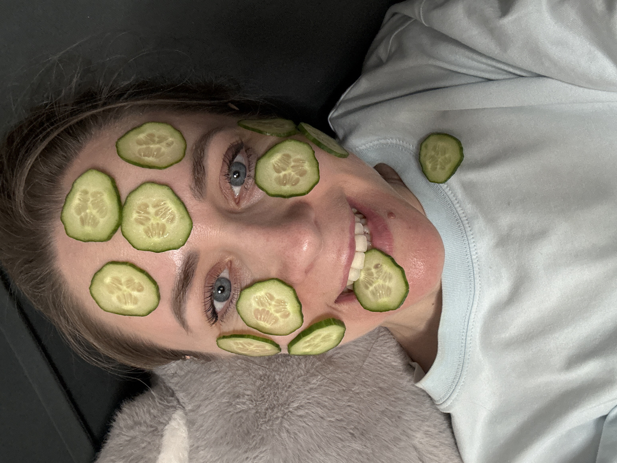 A woman is lying down, wearing a light blue shirt. She has cucumber slices on her face, including her forehead, cheeks, and chin. She is smiling, and her teeth are visible. The background is dark and undefined. The scene does not contain sufficient information to determine if it is located in Chaikovsky, Russia.