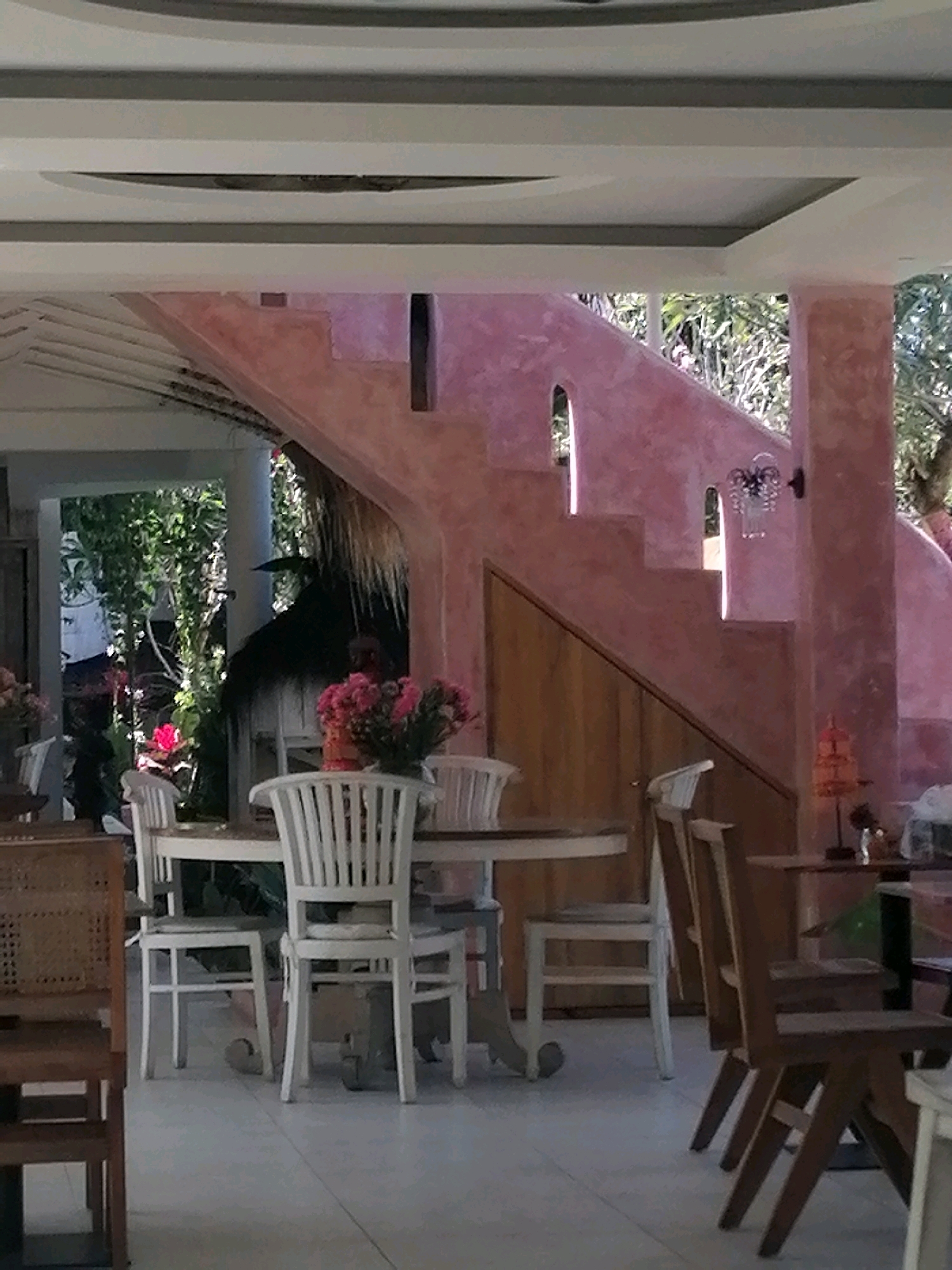 The image shows an outdoor dining area with a pink, curved staircase in the background. Several tables and chairs are arranged around the area. In the foreground, a round table is set with white chairs. A bouquet of pink flowers is placed on the table. To the left, a wicker chair is visible. Behind the pink staircase, lush greenery suggests an outdoor setting. The scene appears to be in a cafe or restaurant, possibly located in the Ubud District of Indonesia, given the tropical vegetation and architectural style.