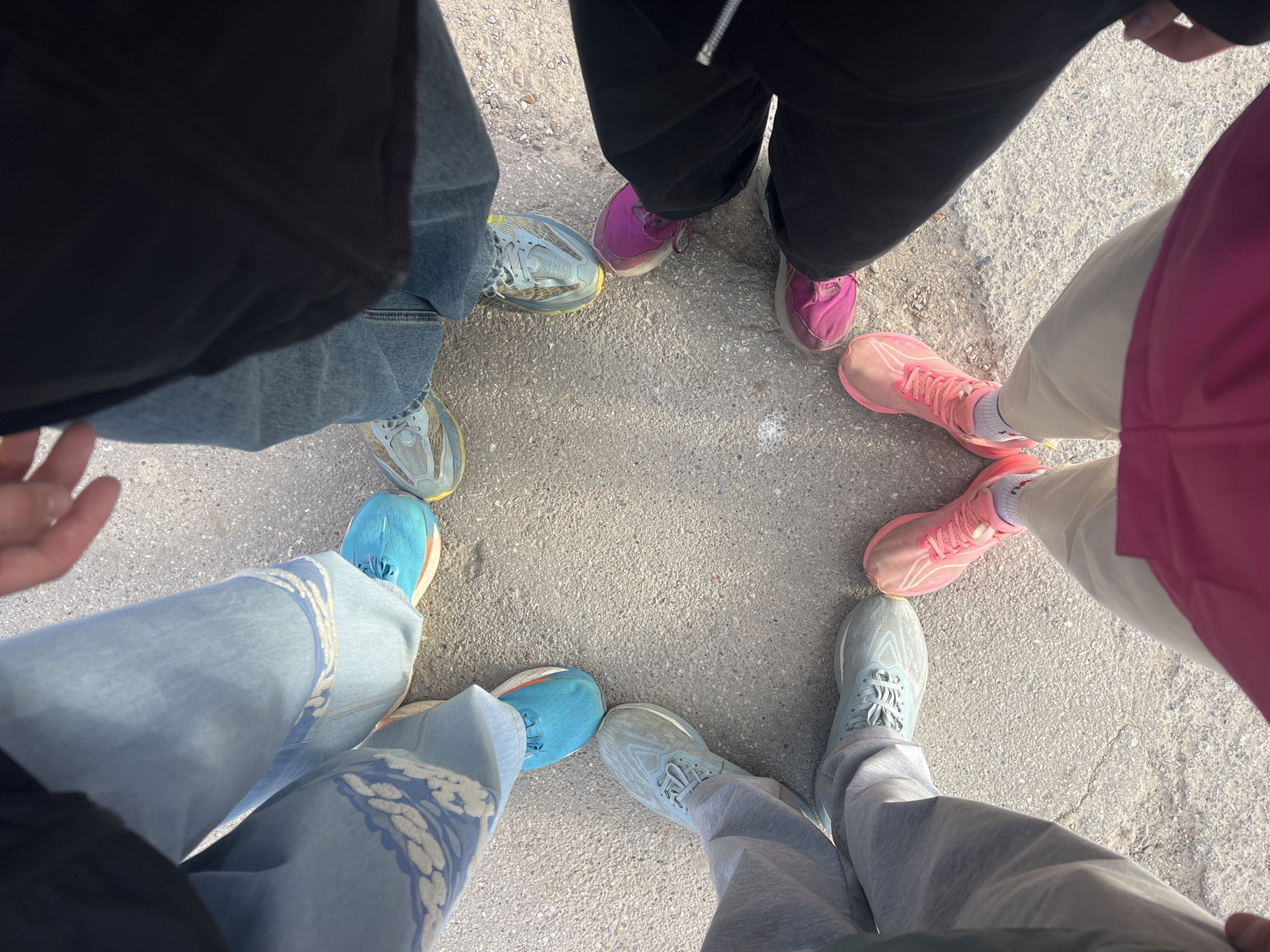 A high-angle shot shows a circle of people standing on a concrete surface. The focus is on their feet and lower legs, revealing different types of footwear and clothing. Two people are wearing blue sneakers paired with light blue jeans, one with a floral pattern. Another person is wearing gray sneakers and gray sweatpants. A fourth person is wearing pink sneakers and light beige pants. There are two people in dark sweatpants and pink or purple sneakers, and another in stonewash jeans and gray sneakers. The image is taken outdoors. The texture of the concrete suggests the location is likely an urban area. There is nothing to indicate this image was taken in Perm, Russia.