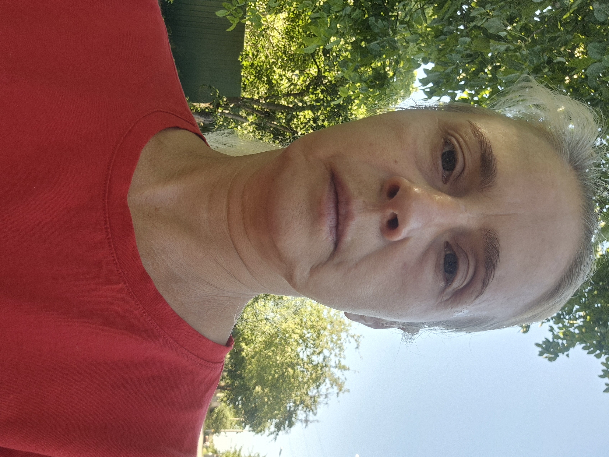 An outdoor, medium shot selfie shows a fair-skinned woman with blonde hair pulled back. She is wearing a red t-shirt. Her face is slightly flushed, and her skin shows signs of aging. The background consists of green trees and a clear blue sky. There is a wooden fence on the left side of the image and a street with trees and utility poles on the right. The location is identified as Sozonivka, Ukraine.