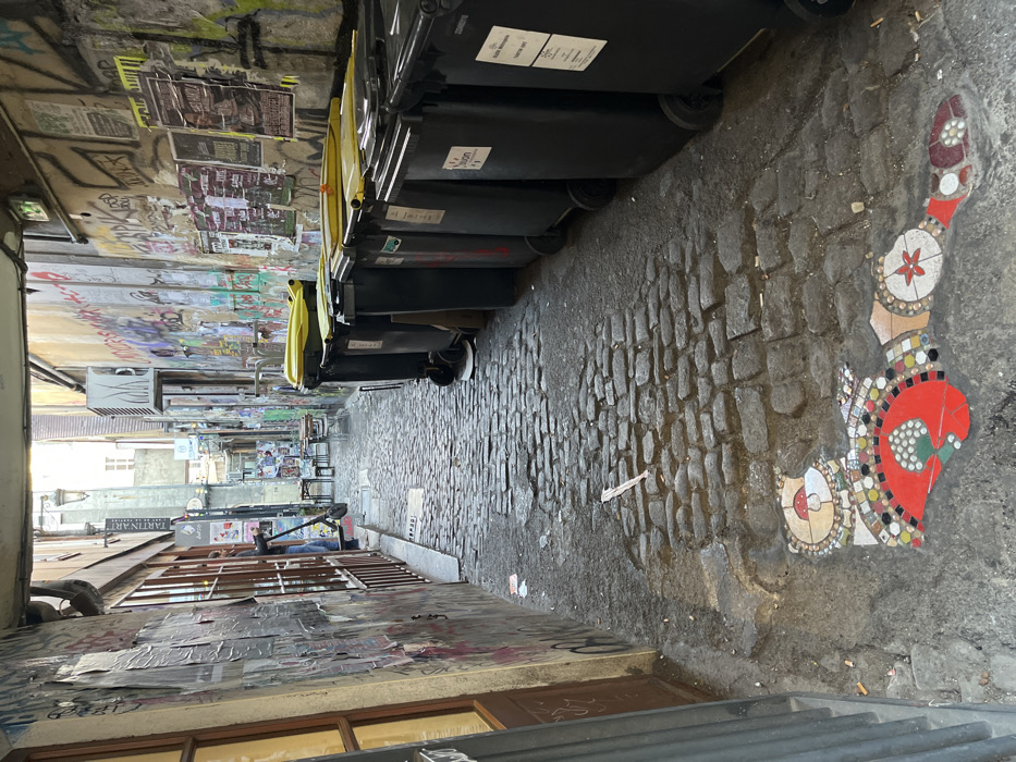 This image captures the Rue des Godrans, a narrow, cobbled pedestrian alleyway in Dijon, France, viewed from its entrance. The ground is paved with irregular cobblestones, showing areas of dampness and scattered debris, including cigarette butts and a piece of paper. Near the bottom left, an embedded mosaic composed of various colored tiles forms a stylized, elongated figure on the cobblestones.
The left wall of the alley is densely covered with layered posters, flyers, and graffiti. A dark brown wooden door or window frame with glass panes occupies the immediate left foreground. Further into the alley on the left, a dark-colored electric scooter is parked, partially obscured by an individual visible from the waist down, wearing blue jeans. A sign labeled "TARTIN'ART" is visible further down this wall.
Along the right side of the alley, a continuous line of approximately eight dark gray wheeled waste bins with yellow lids is positioned against the wall, extending into the midground. The right wall is also extensively covered with graffiti, layered posters, and some areas of green paint. An air conditioning unit is mounted high on this wall in the midground.
In the midground, the alley narrows and curves slightly to the left. Several small, round tables with matching chairs are arranged on the cobblestones, indicating an outdoor seating area. The far end of the alley opens into a brighter urban space, revealing a taller building and a distant street lamp, under natural daylight conditions.