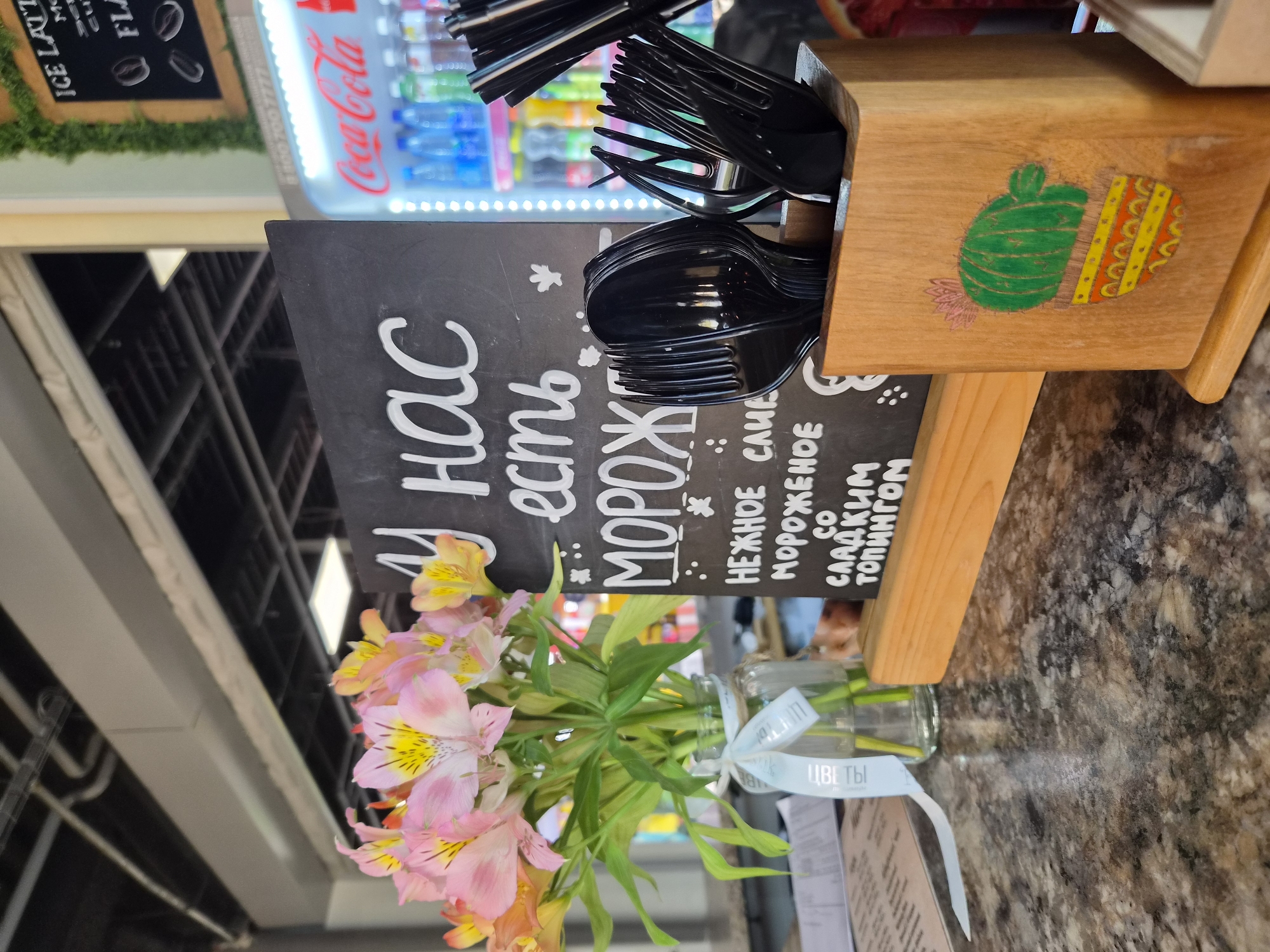 This is a medium-shot, eye-level, indoor photograph of a counter at a cafe or similar establishment, likely in Arseniev, Russia. The focus is on a chalkboard sign and a bouquet of flowers.
On the left, a bouquet of light pink and yellow alstroemeria flowers is arranged in a clear glass vase. A white ribbon with Russian text that appears to read "ЦВЕТЫ" (flowers) is tied around the neck of the vase. The flowers are in the foreground and are slightly blurred due to shallow depth of field.
To the right of the flowers, a small, black chalkboard is propped up on a wooden stand. White chalk lettering on the board reads, "У нас есть МОРОЖНОЕ" (We have ICE CREAM). Below this, it lists "НЕЖНОЕ САМЕ МОРОЖЕНОЕ СО СЛАДКИМ ТОПИНГОМ" (DELICATE ICE CREAM WITH SWEET TOPPING).
In front of the chalkboard, to the right, is a wooden holder containing black plastic forks and spoons. The wooden holder has a drawing of a green cactus in a colorful pot.
In the background, a brightly lit Coca-Cola refrigerator is visible, stocked with bottles and cans. Above the counter, industrial-style lighting and conduits are present. The overall environment suggests a casual, possibly fast-food or cafe setting. No people are visible in the frame.