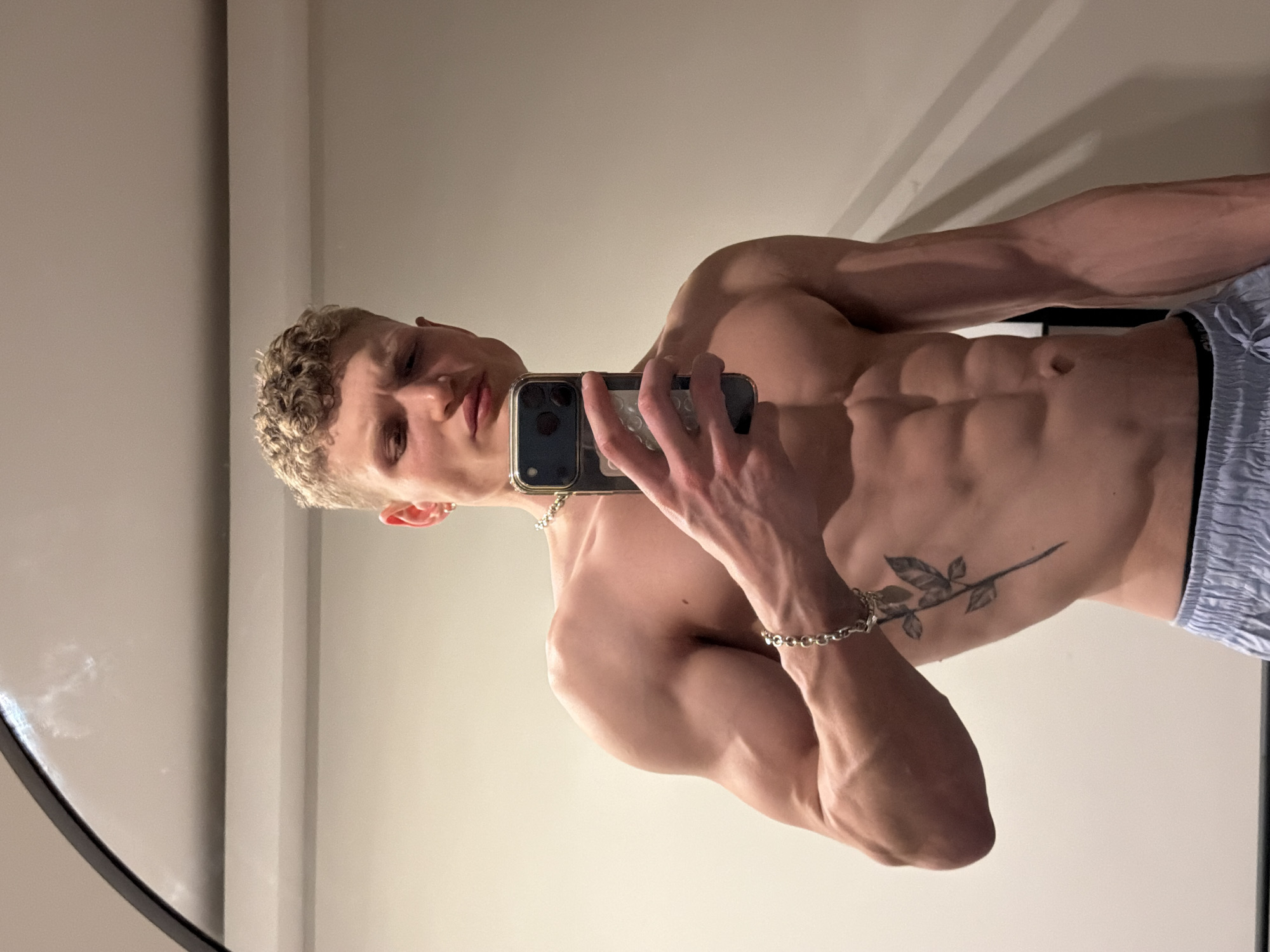 A shirtless, light-skinned male with blonde curly hair is taking a selfie with a smartphone. He is wearing light blue patterned shorts, a silver necklace, and a bracelet on his left wrist. He has a tattoo of a branch with leaves on his left side. He stands indoors against a neutral colored wall. A partial view of a mirror is visible on the left side of the frame. The location cannot be determined from the image.