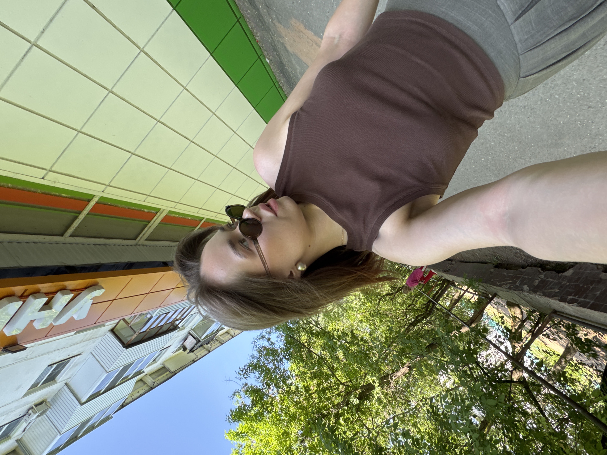 A full-length, low-angle, personal photograph depicts a female in Chaikovsky, Russia, standing outdoors. The subject wears a brown tank top and grey shorts. She has sunglasses and a single visible earring. The background includes a building with a sign (partially visible), a blue sky, and green trees. A tiled wall with white, green, and orange sections is also in the background. The ground appears to be concrete.