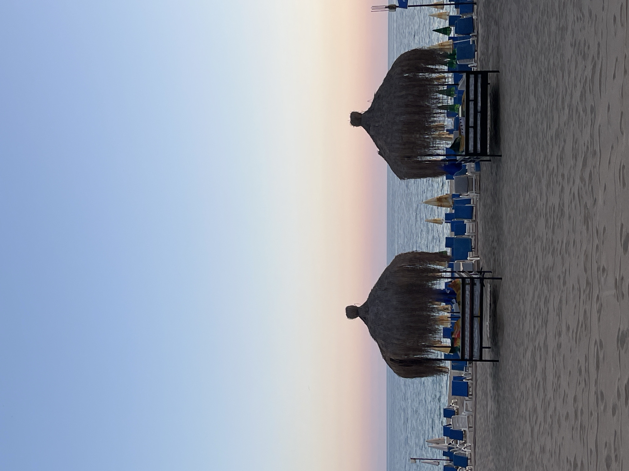 This image captures a tranquil beach scene, likely at sunrise or sunset, on a clear day in Alanya, Turkey. The sky, occupying the upper portion of the frame, displays a gentle gradient, transitioning from a soft, light blue on the left to delicate hues of pale pink and orange near the horizon on the right. Below the expansive sky, the calm, placid sea stretches across the midground, reflecting the subdued light.
The foreground is dominated by the sandy beach, showing subtle textures and scattered footprints. Two large, dark-thatched beach umbrellas, or palapas, stand prominently on the sand closer to the water's edge, their conical shapes silhouetted against the lighter background. Behind and around these main structures, an array of sun loungers is neatly arranged, most of them blue, with some white ones interspersed. Several smaller, conventional beach umbrellas, primarily blue, yellow/beige, and a few green, provide additional shade.
The beach appears devoid of people, suggesting a quiet moment either before the day's activities commence or after they have concluded, enhancing the sense of peace and serenity. No specific activities are taking place, and the scene exudes a calm atmosphere. There is no visible text in the image.