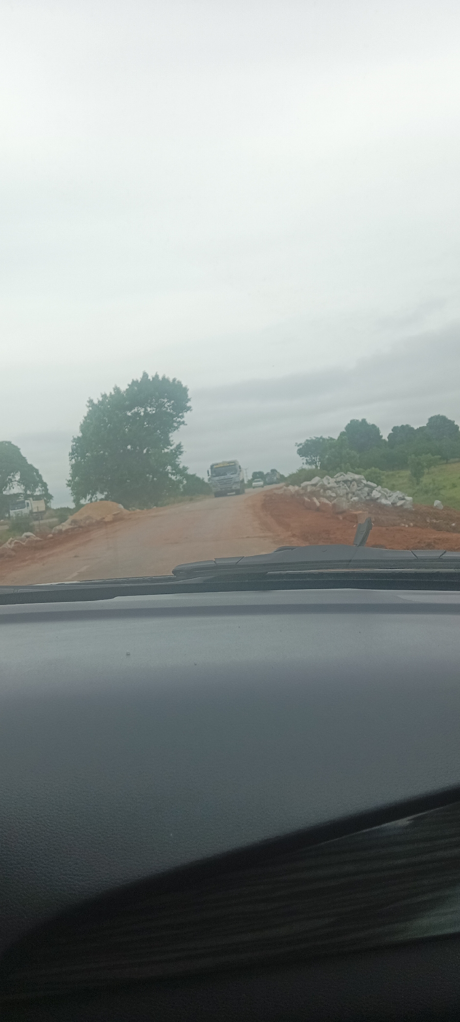 The image shows a view from inside a vehicle traveling on a dirt road in Kadarko, Nigeria. The road appears to be under construction, with exposed reddish-brown soil on the sides. A large pile of rocks or rubble sits adjacent to the road on the right side. In the distance, several vehicles are visible, including a truck, which could indicate ongoing construction activity. The sky is overcast, creating a subdued, gray ambiance. In the foreground, the vehicle's dashboard and windshield wipers are in focus, while the rest of the scene is further away. The environment consists of trees and grassy land visible beyond the road, typical of a rural or semi-rural setting. No people are directly visible in the image.