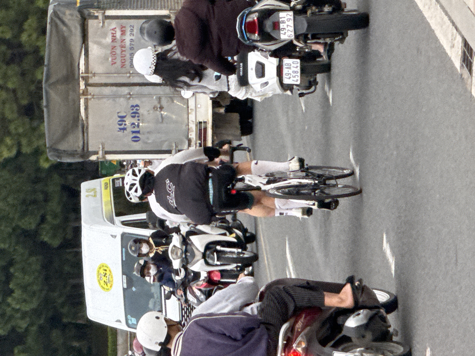 The image depicts a street scene in Da Lat, Vietnam, with several vehicles and people on the road. In the foreground, a person on a red scooter is visible from the back. To the left, a white van with "2.8si" and "2S" written on it is parked. In the center, a person on a bicycle with a black jacket and white helmet is cycling. Behind the cyclist, a white truck with Vietnamese writing, including "VƯỜN NHÀ NGUYỄN MY 0905 579 292", is prominent. Further back, several motorcycles are seen, some carrying riders wearing helmets and masks. The road surface is asphalt, with white lane markings. Trees line the background, suggesting an urban or suburban setting. The overall impression is a busy street with typical traffic conditions.