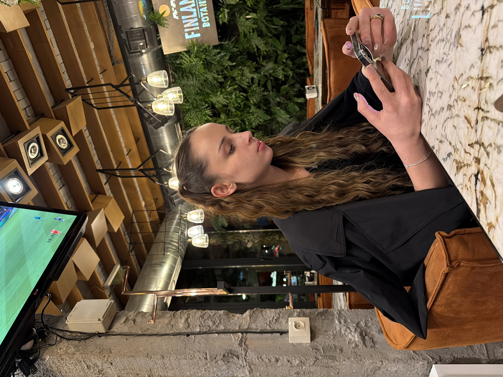 This image captures a young woman seated in a modern restaurant or bar in Sofia, Bulgaria, during the evening. The primary subject, shown in profile, has long, wavy brown hair and is wearing a black top or jacket. She is focused downwards, holding a small metallic object, possibly a vape or a similar device, in her manicured hands. A ring is visible on her right hand, and a delicate bracelet adorns her left wrist.
She is seated at a table with a light-colored, patterned surface that resembles marble or granite, and rests on a brown upholstered chair or booth. In the background, a large television screen displays a live soccer or football match, showing a green field, players, a "2:0" score, and an "88:34" timer. The "LG" logo is also visible on the TV.
The establishment's interior features an industrial-chic design. Wooden slatted elements form part of the ceiling or upper walls, complementing the exposed grey brick or stone walls. Modern pendant lights with clear glass shades illuminate the space. Behind the woman, a wall is adorned with green foliage, possibly artificial plants. Above these plants, a sign with partially visible text reads "FINLAND BOTANICAL." Metal pipes or ducts are visible overhead. The overall ambiance suggests a casual and contemporary setting, suitable for dining or socializing while watching sports.