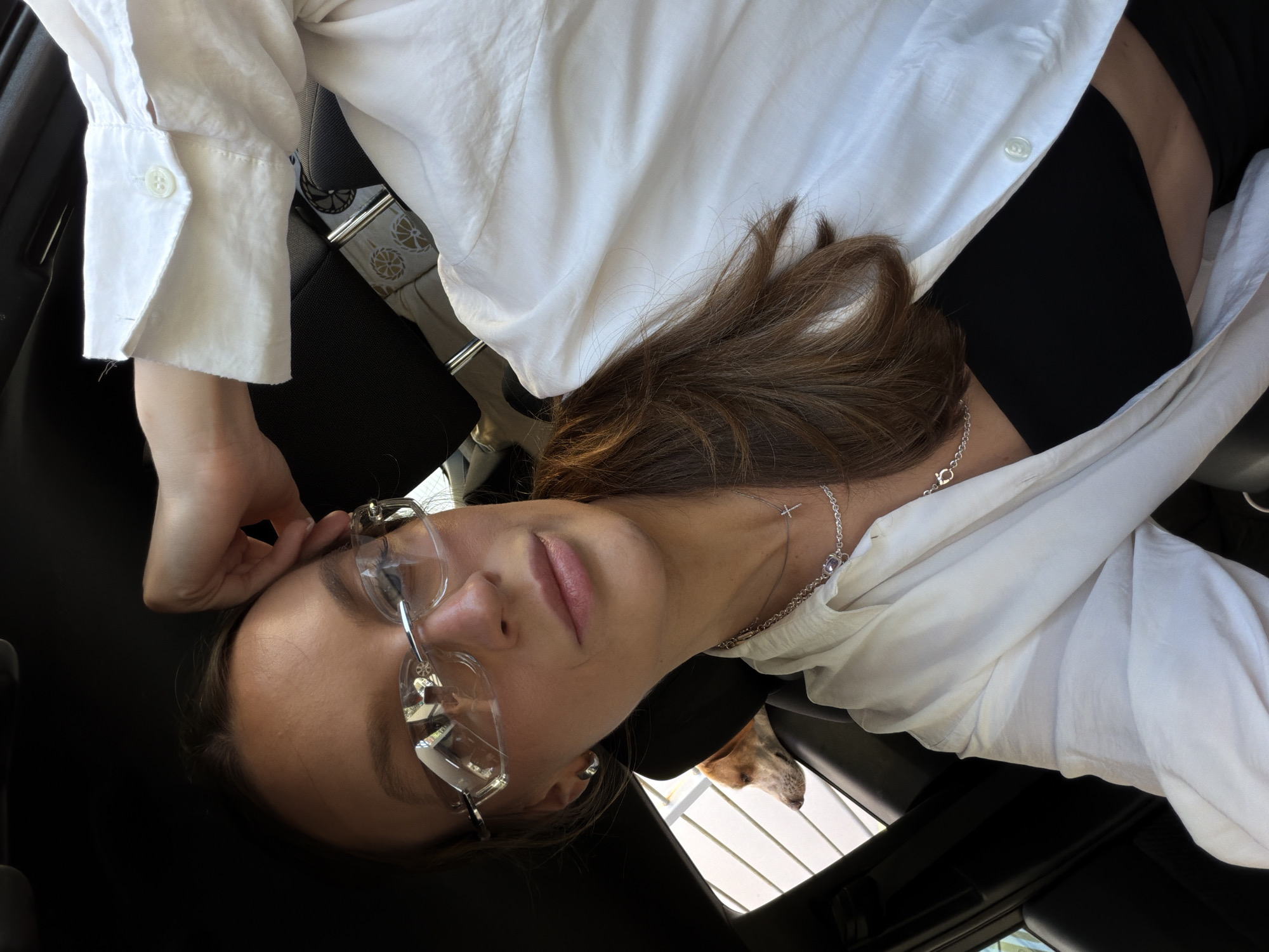 A young woman with long brown hair and glasses is sitting in the driver's seat of a car. She is wearing a white button-up shirt over a black top and a silver necklace. Her right hand is resting on her head. A dog is visible in the back seat, looking out the window. The car appears to be parked outdoors, with a building visible through the window. The lighting suggests it is daytime. This scene is set in Budva, Montenegro.