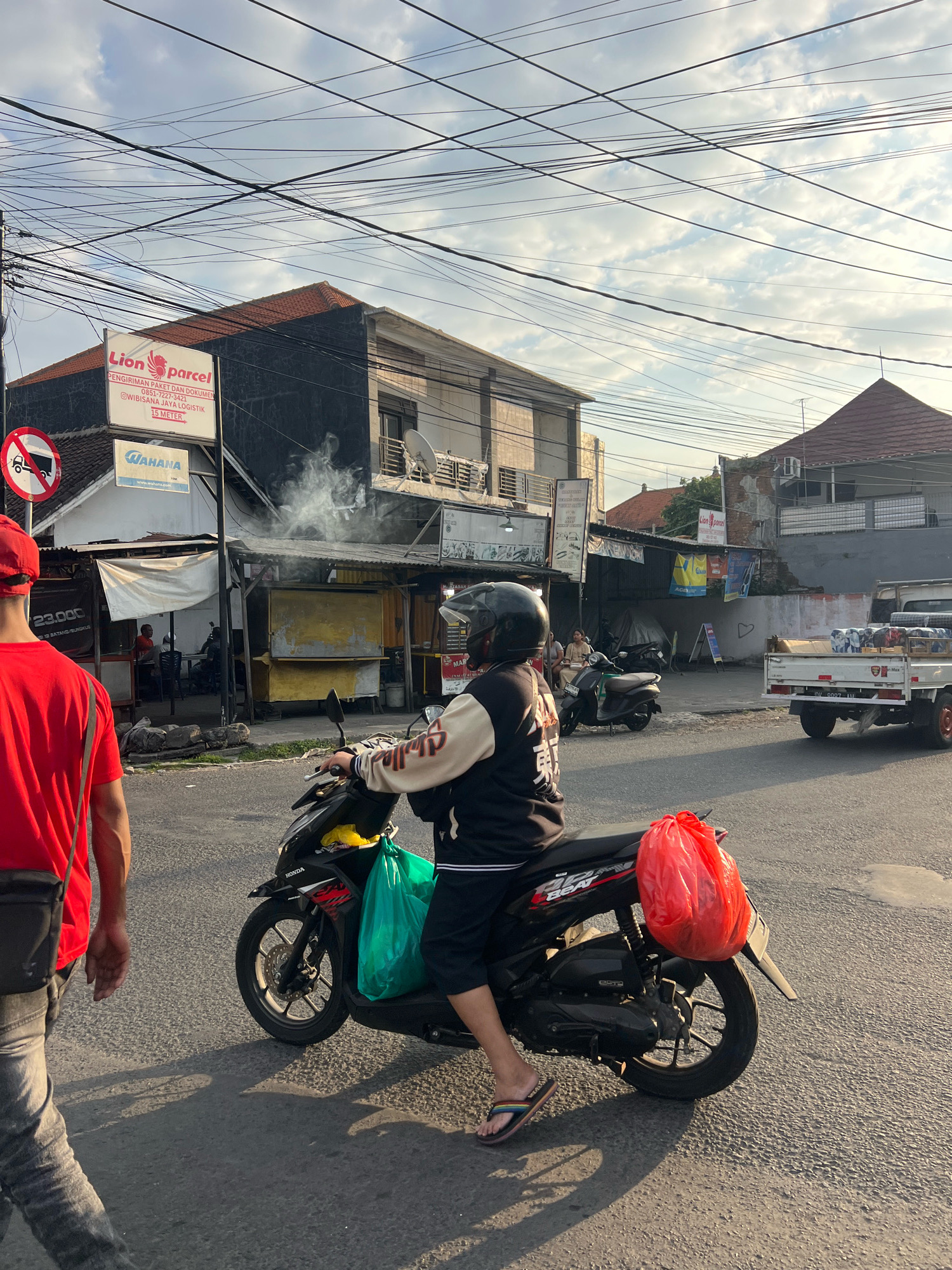 In Denpasar, Indonesia, on a street with overhead wires, a person wearing a black helmet and a black and cream jacket rides a black scooter. The scooter has a bright orange bag attached to the rear and a green bag hanging on the left side. To the left, a person in a red shirt and grey pants, with a black sling bag, walks away from the camera. Behind the scooter rider, a white truck with a load of blue-wrapped items drives away. In the background, buildings line the street, including one with a "Lion Parcel" sign and a "Wahana" sign. Smoke is rising from a stall in front of a building. The sun is casting long shadows on the asphalt road.