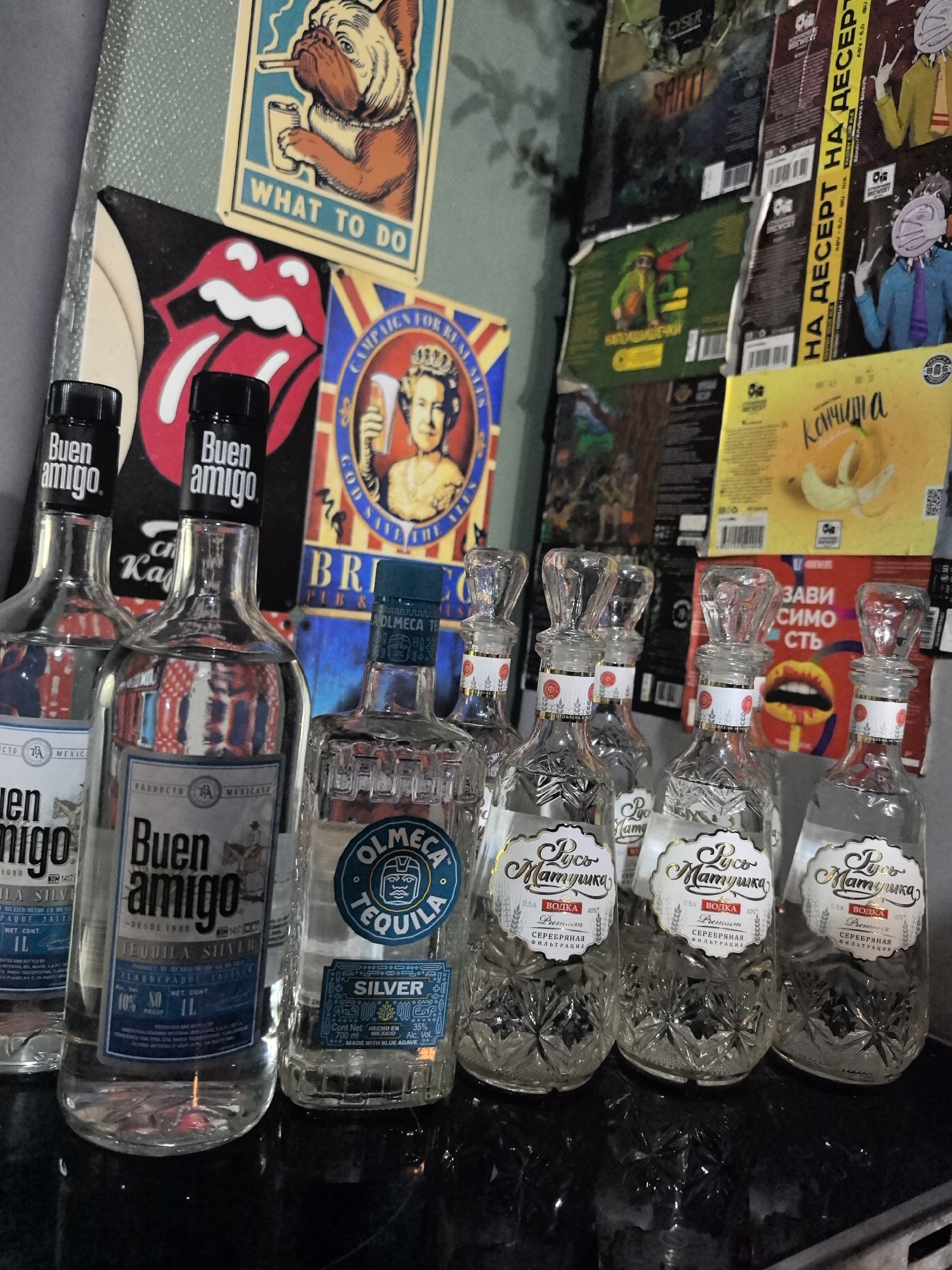 The image presents a variety of alcoholic beverages on a dark surface, possibly a bar counter, in front of a wall decorated with an assortment of posters and labels. Two bottles of "Buen Amigo" tequila are positioned to the left, followed by a bottle of "Olmeca Tequila Silver." To the right are four bottles labeled "Русь Матушка" vodka, each with ornate crystal-like glass.
The backdrop consists of an eclectic mix of promotional materials. There is a cartoonish poster of a bulldog smoking and holding a beverage, a "Rolling Stones" tongue logo, a "Campaigns for Real Ales" poster featuring Queen Elizabeth II, and various promotional labels, including one with the text "НА ДЕСЕРТ НА ДЕСЕРТ." Another label shows the name "Кончита" with banana images, and yet another shows a pair of lips.
Based on the Cyrillic lettering on the vodka bottles ("Русь Матушка"), this image appears to be taken in Russia, specifically Novi.