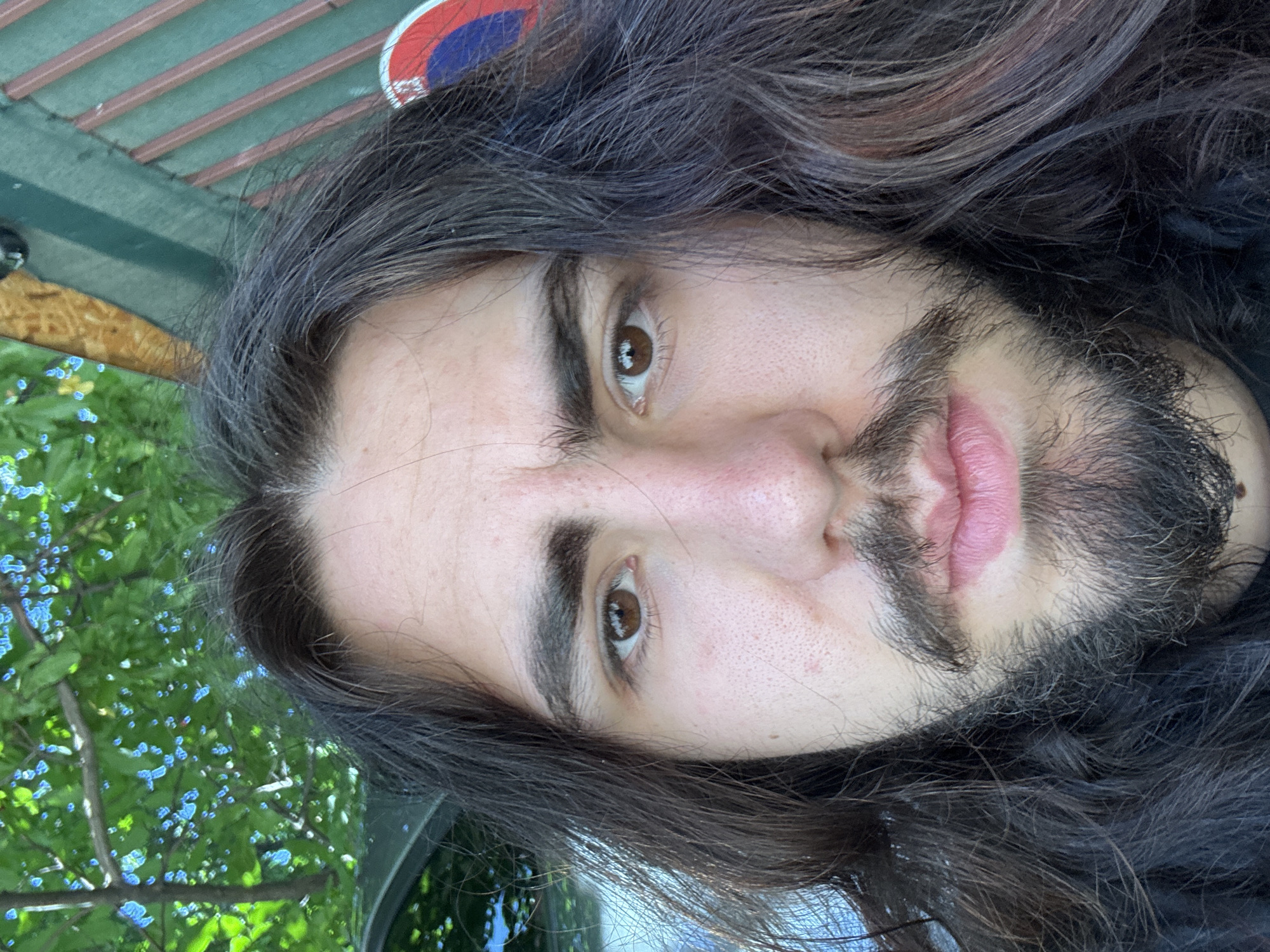 An extreme close-up selfie of a person with long, dark, wavy hair and a beard. The person has brown eyes and fair skin. The background is out of focus and shows green foliage and a hint of a dark green and red striped structure. There is also a partially visible circular sign with red and blue elements. The lighting suggests it is daytime.