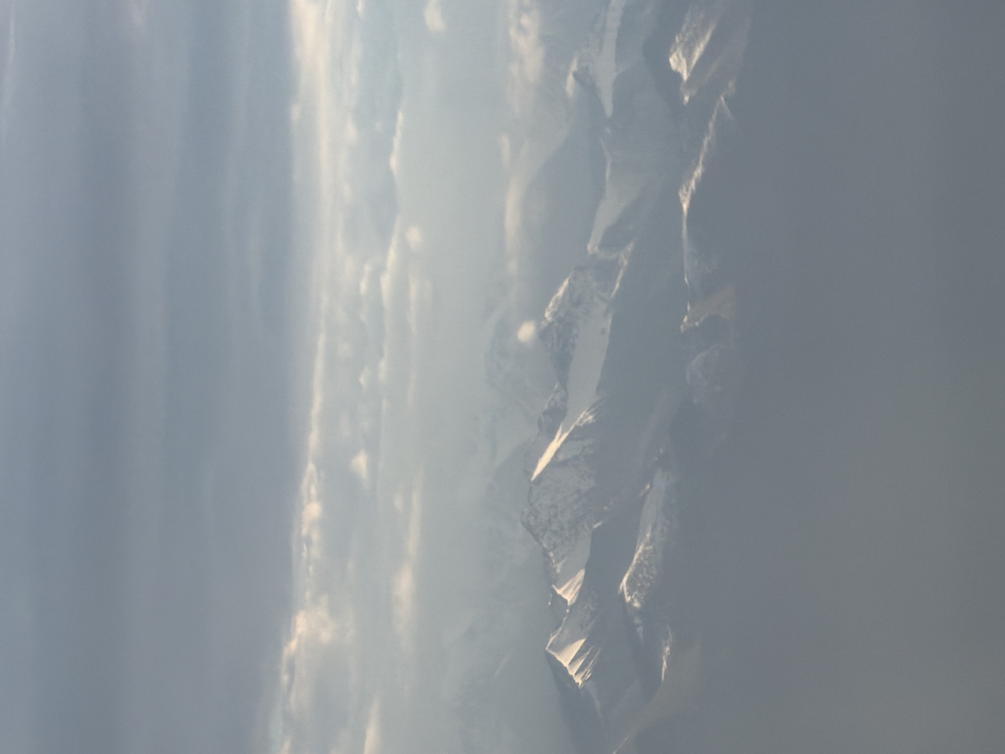 The image shows a vast expanse of snow-capped mountains, likely in the Himalayan region. The sky above is filled with clouds, some dark and ominous, others illuminated by the sun, creating a dramatic contrast. The light filtering through the clouds casts a soft glow on the mountain peaks, highlighting their rugged contours. The lower part of the image is obscured by a layer of mist or haze, adding a sense of depth and mystery to the scene. The overall impression is one of grandeur and remoteness, evoking the majesty of nature in its rawest form.