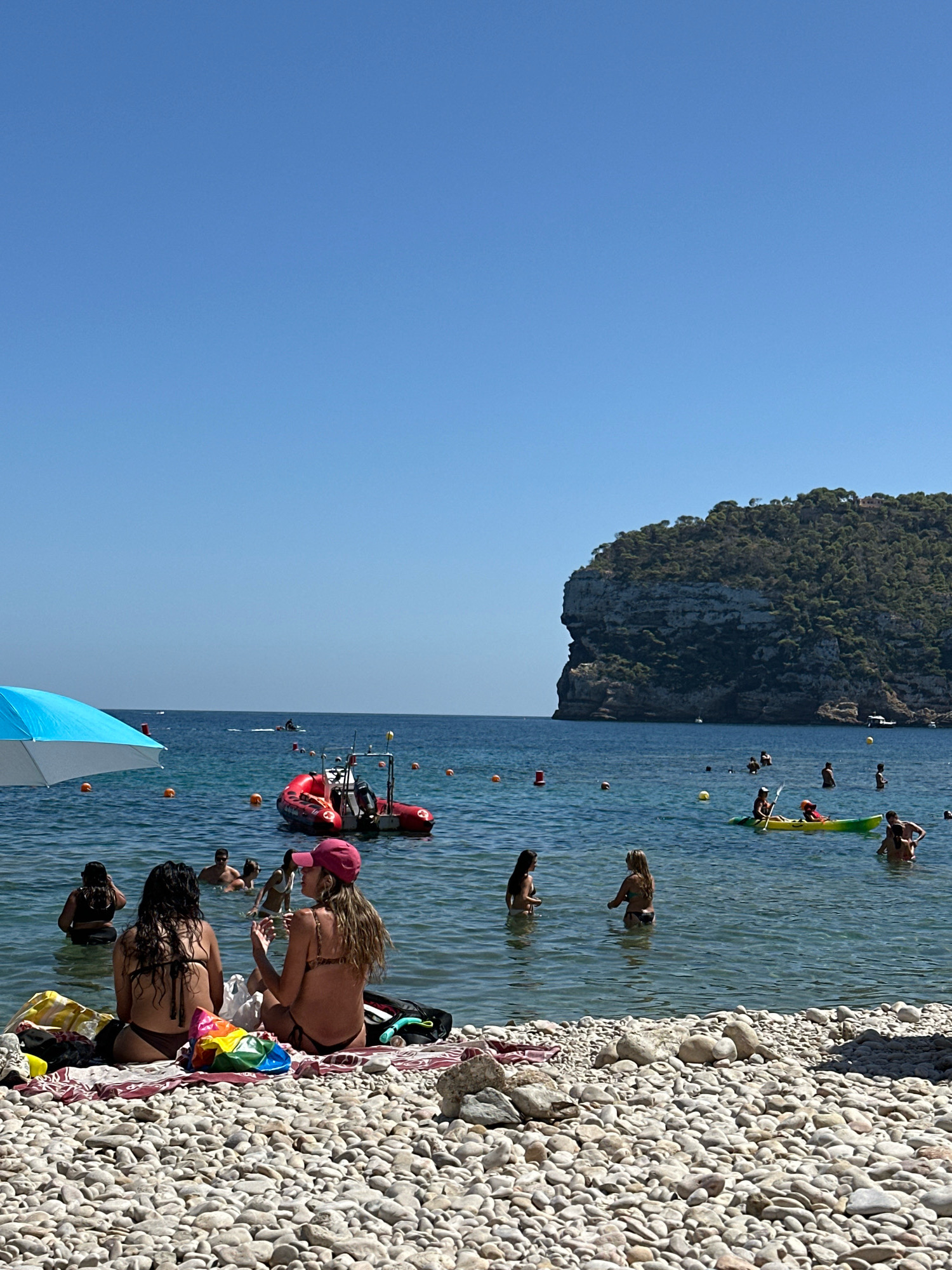 This is a photograph taken on a sunny day at a pebble beach in Xàbia, Spain. In the foreground, two women are sitting on beach towels with their backs to the camera, facing the water. They are wearing bikinis. The beach is covered in smooth, white pebbles. To the left, a blue and white beach umbrella is partially visible. In the shallow, clear blue water, several people are swimming, wading, and sitting. Further out in the sea, a red inflatable boat with a person standing in it is visible. Another small boat is further in the distance. A person is paddling a yellow kayak. In the background, a large, tree-covered rocky headland rises from the sea, characteristic of the coastline of Xàbia. The sky is a clear, bright blue.