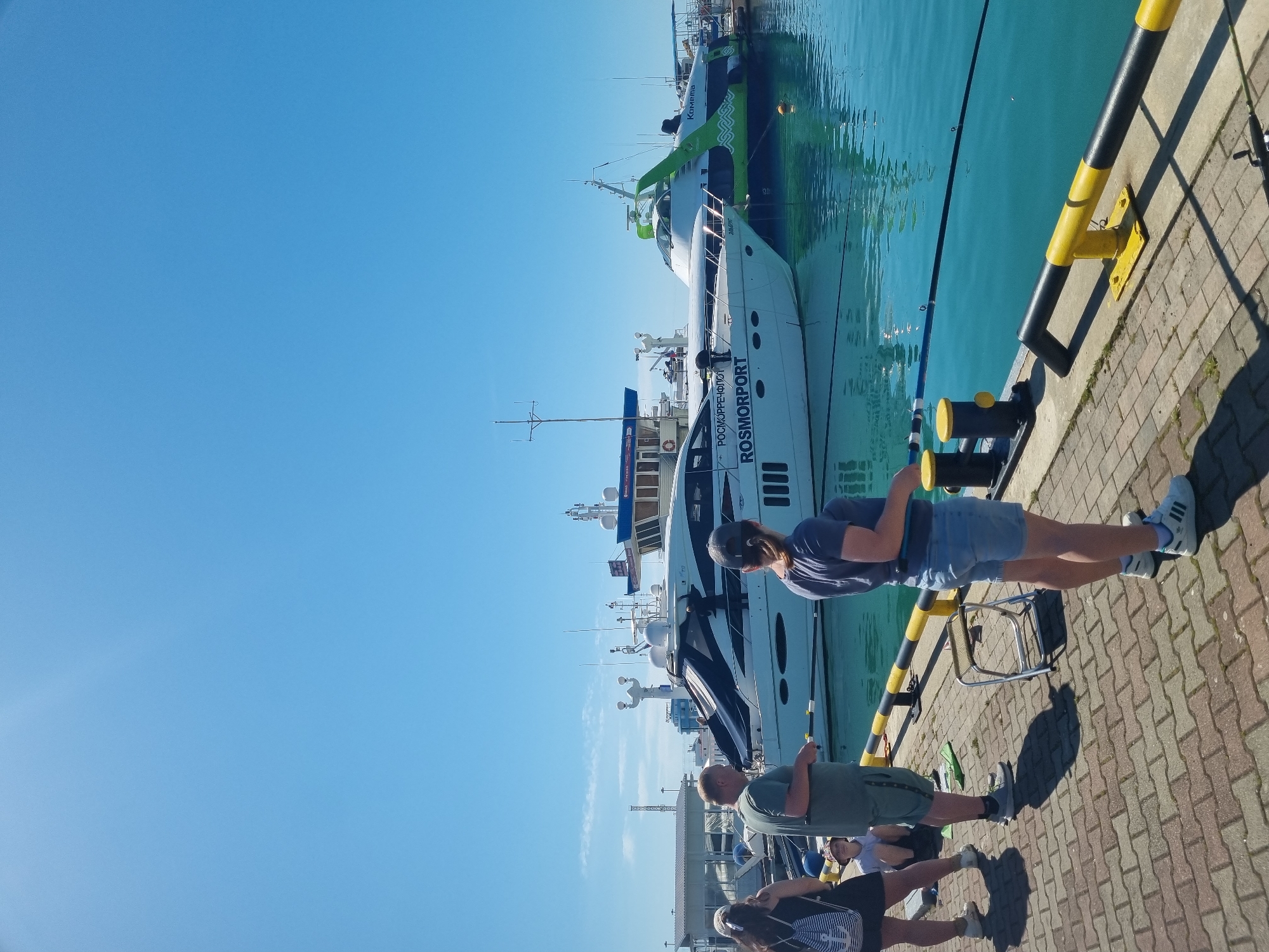The image depicts a sunny day at a harbor, likely in Sochi, Russia, given the presence of a vessel with "ROSMORPORT" inscribed on its side, which is the name of a Russian state company dealing with port infrastructure. A group of people are seen standing on the brick-paved harbor, next to a black and yellow bollard. One person, dressed in a blue shirt and denim shorts, is holding a fishing rod, suggesting they are fishing. A small, metal folding chair is nearby. A large, white passenger vessel, possibly a hydrofoil, is docked in the background. The sea appears calm and turquoise in color. The sky is a clear, bright blue.