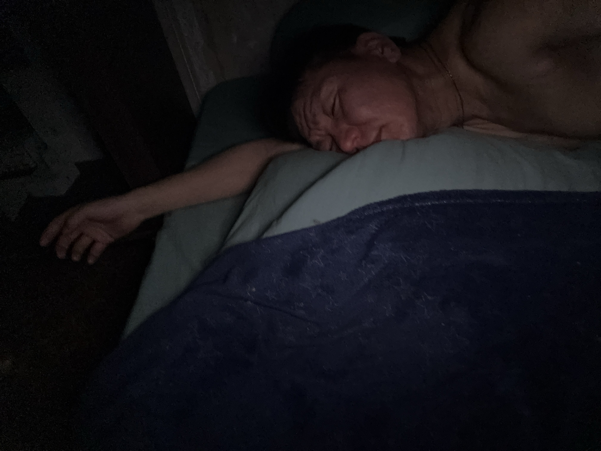 The image shows an older man sleeping in a bed. He is lying on his side, with his head on a light green pillow. A dark blue blanket covers part of his body. His left arm is extended to the side. The scene is dimly lit. The location is unknown and cannot be determined from the image.