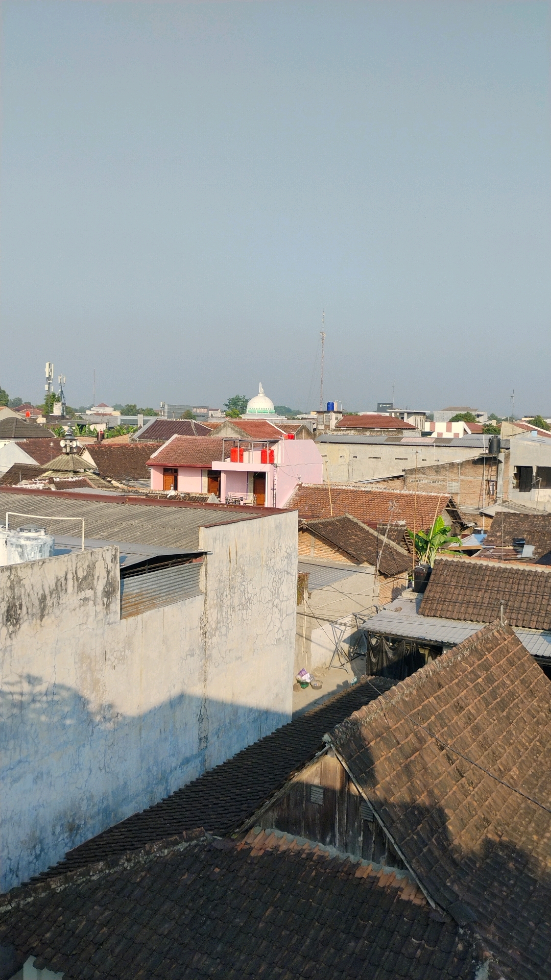 This is an aerial, eye-level, full shot of rooftops in Gumpang, Indonesia. The image captures a dense residential area with various buildings and structures. The sky is clear and pale blue, indicating it is daytime.
In the foreground, several rooftops are visible. The closest are covered with dark, weathered tiles, some appearing worn. A light-colored, textured wall dominates the left side of the foreground, casting a long shadow across a tiled roof.
Further into the scene, more rooftops of varying colors and materials can be seen. A notable pink building with white trim and red water tanks on its roof stands out among the predominantly brown and grey roofs. Behind this pink building, a white dome, likely part of a mosque or similar structure, is visible in the distance, topped with a spire.
Various antennas and communication towers are scattered across the skyline, suggesting a connection to infrastructure. The overall impression is of a typical urban or semi-urban residential landscape in Indonesia. No people are clearly visible in the image. The lighting suggests it might be morning or late afternoon due to the length of the shadows.